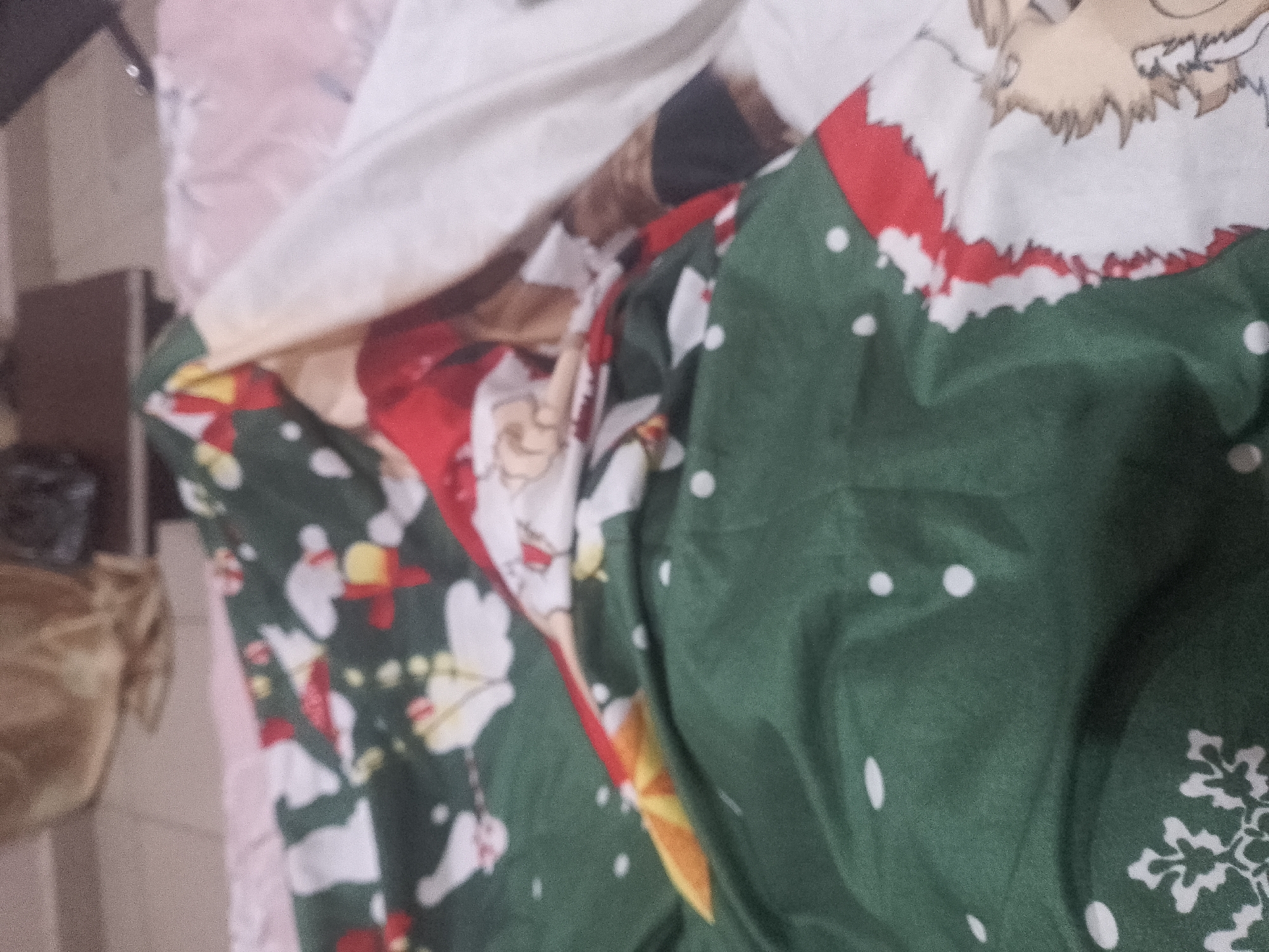A close-up, angled shot shows a pile of fabric with a Christmas-themed pattern. The dominant colors are green, red, and white. The fabric appears to be a duvet cover or sheet, featuring depictions of Santa Claus, snowflakes, and scattered white dots. The material is creased and folded, creating shadows and highlights that obscure some of the details. In the background, a portion of a pale pink fabric is visible, as well as a dark, rectangular object and a blurred golden object. The scene is indoors, but the specific location within a city or any environmental context is not discernible from the image.