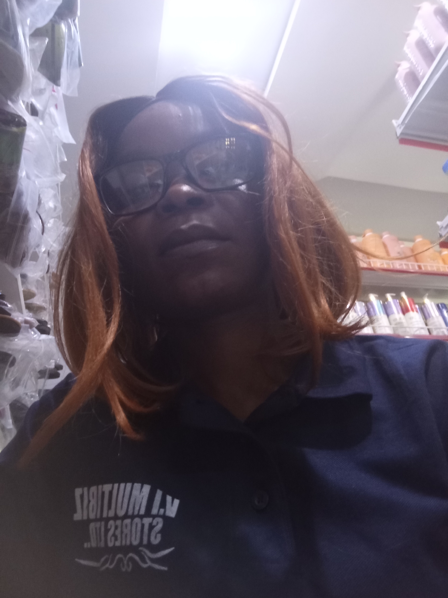 The image shows a close-up, downward-facing selfie of a woman with reddish-brown hair and glasses. She is wearing a dark blue collared shirt with white lettering that appears to be reversed, possibly indicating it is a reflection or a mirrored print. The lettering reads "VI MULTITUM I.V" and below it, ".III 23R0T2 STORES". The background suggests an indoor environment, possibly a store, with shelves lined with various products, some in clear plastic packaging and others in colored bottles. The lighting is bright and overhead. The location is identified as Howayi, Nigeria.