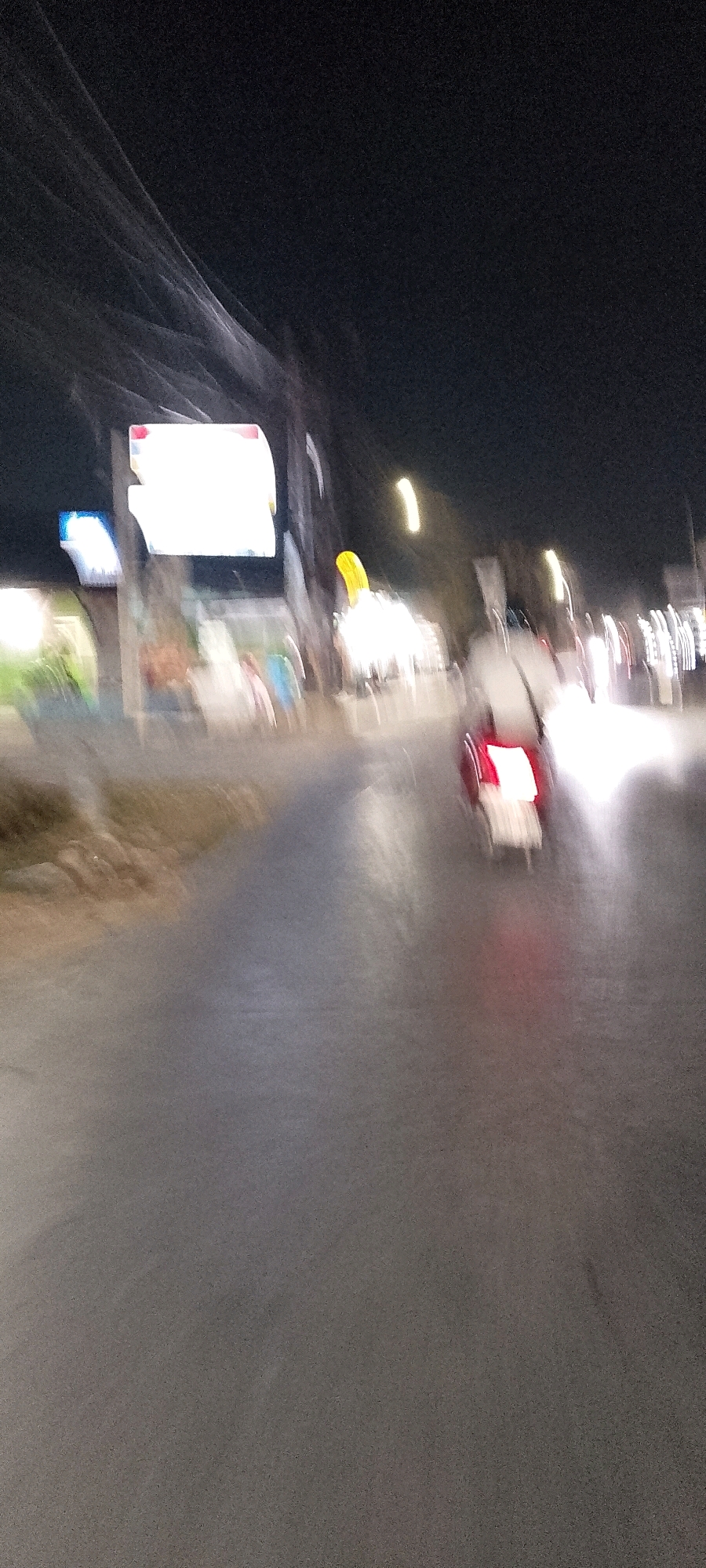 The image depicts a night scene on a street in Satriajaya, Indonesia. The foreground shows a grey asphalt road with some motion blur, suggesting movement. In the middle ground, a red and white vehicle, possibly a scooter or motorcycle, is illuminated by its taillights, which are a bright red. The vehicle is also blurred due to motion. To the left of the road, there are some out-of-focus figures, suggesting pedestrians, and blurred vegetation or embankment. Further back, a bright rectangular sign with red and white elements is visible, likely an advertisement or informational display. Other indistinct lights and blurred shapes are scattered throughout the background, consistent with a nighttime urban environment. Overhead, blurred lines suggest electrical wires or cables.