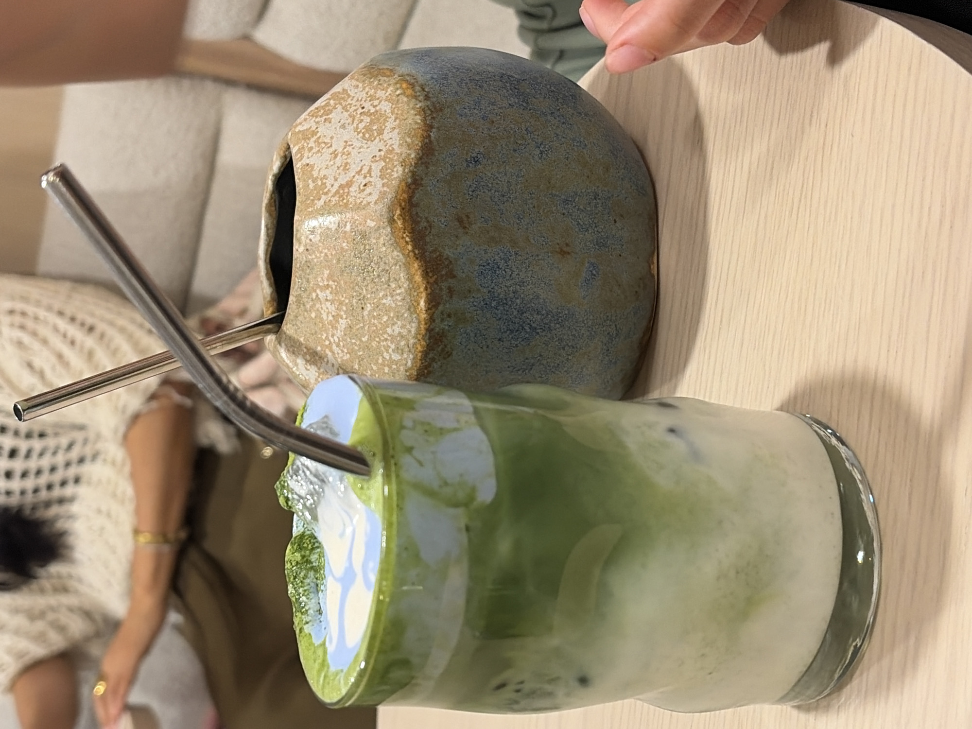 A table holds a light-colored wooden table top. A rounded, porous, ceramic object with light brown and blue hues sits next to a glass filled with a green and white beverage, likely matcha latte, featuring a metal straw. A person's hand and part of their arm is visible at the top of the frame. Another person wearing an off-white knitted top is partially visible, with an arm showing a gold bracelet and a ring. A portion of a white patterned couch can also be seen. Based on the ceramic object and beverage commonly associated with the city, it is likely this image was taken in Puebla, Mexico.