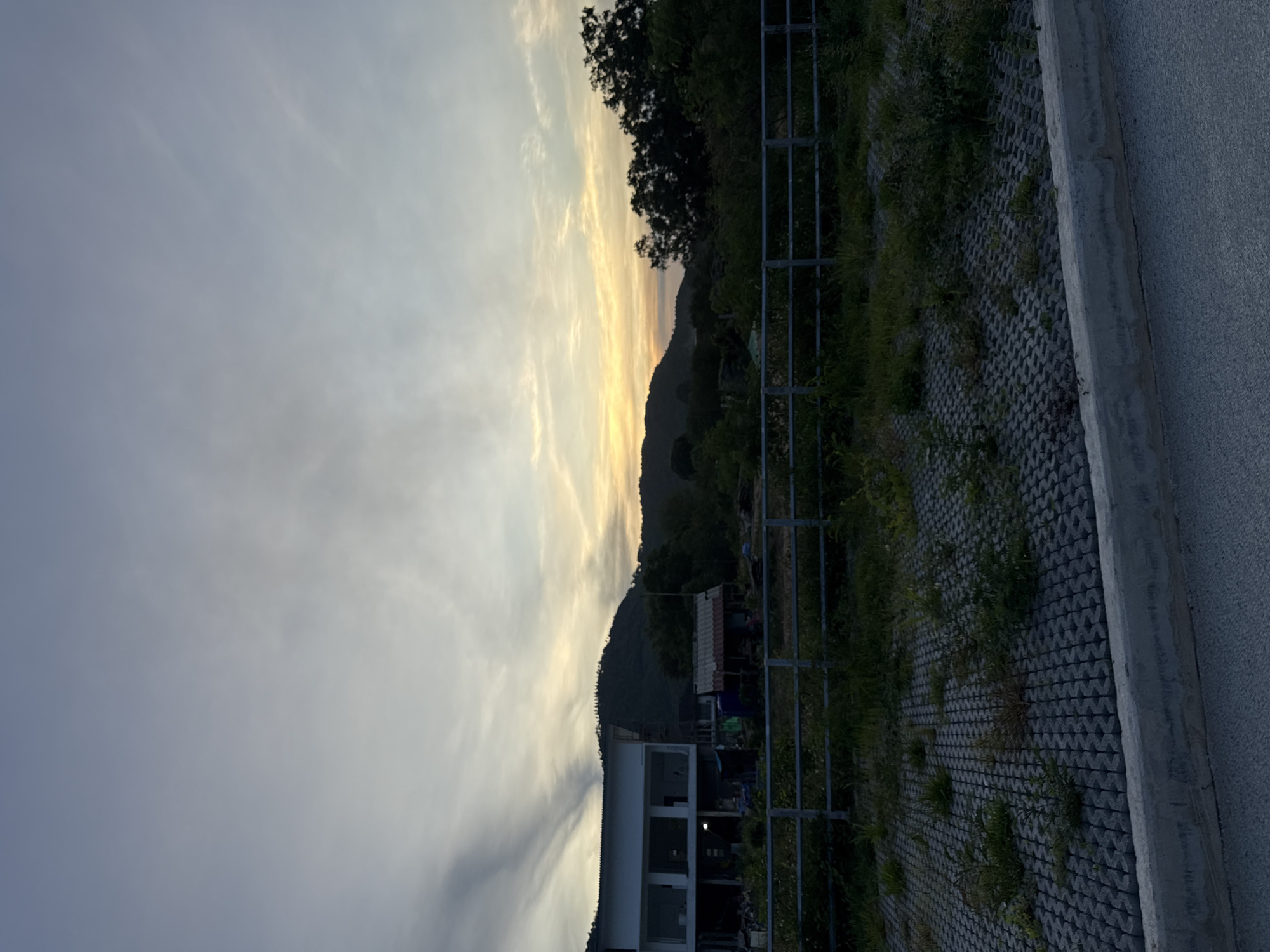The image depicts a landscape scene at dusk with a wide sky filled with streaks of white and grey clouds, transitioning to yellow and orange hues near the horizon. In the foreground, a textured grey pavement forms a border with a concrete curb. Beyond this, a gravelly area is visible with patches of green grass and weeds growing through it, separated by a metal railing. In the mid-ground, a building with a white facade and multiple windows is under construction or renovation. Adjacent to it is a smaller structure with a red and white striped roof. Behind these buildings, a dense line of green trees and foliage is present, with a large, dark green hill or mountain rising behind them. The overall setting appears to be semi-urban or rural, possibly on the outskirts of a town. The location is identified as Baan Bang Rak, Thailand. No people are visible in the image.