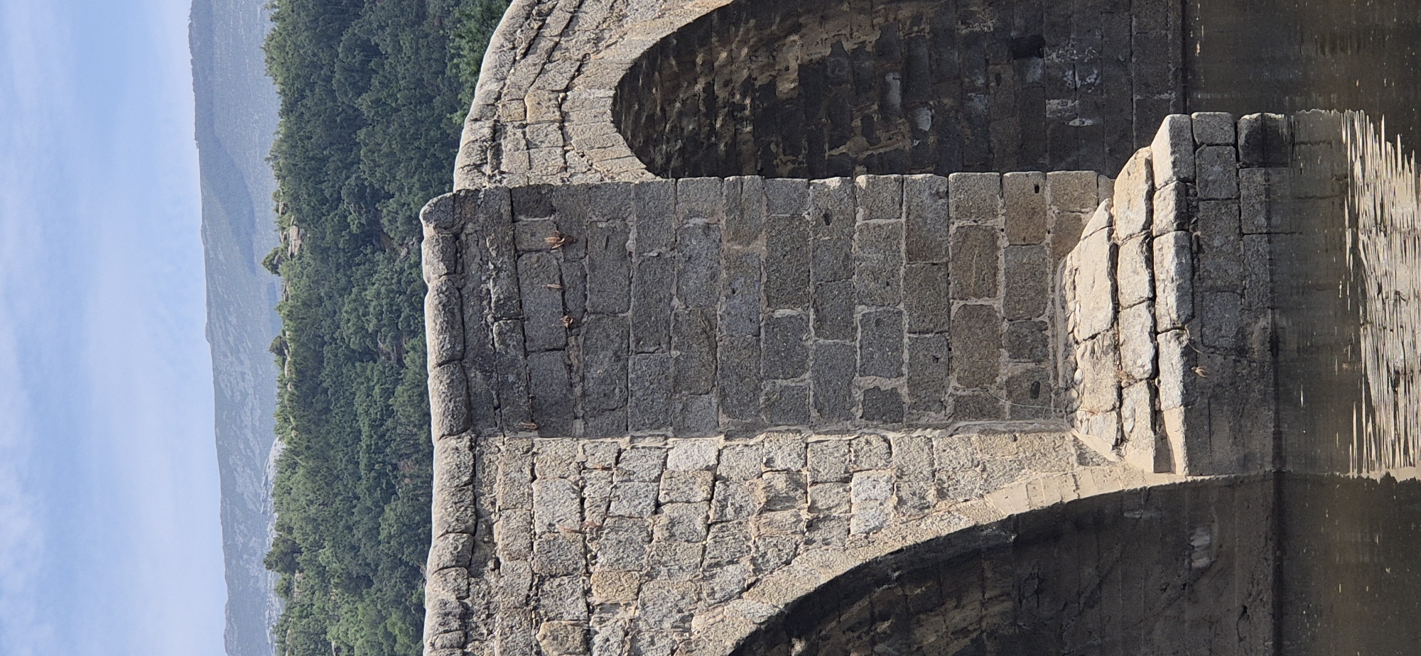 The image shows a close-up view of an old stone bridge arch, with a lush green forest and a mountain range in the background under a clear blue sky. The bridge is constructed from rough-hewn gray stones, varying in size and shape, laid in courses. Some sections of the wall are topped with rounded stones, suggesting the arch's form. The texture of the stone is rough and weathered, indicating its age.
Beneath the bridge, the water's surface is visible, reflecting the stone structure in a distorted manner. The reflection creates shimmering vertical lines, suggesting movement in the water. The lighting indicates it is daytime, likely with the sun casting shadows that highlight the stone's contours.
In the distance, a dense forest covers the slopes of a mountain. The trees are a vibrant green, and the mountain itself is a hazy blue-gray, suggesting a considerable distance and atmospheric perspective. The sky is a bright, cloudless blue, contributing to the impression of a clear and sunny day.
No people are visible in the image. The focus is entirely on the architectural structure and the natural landscape. The overall impression is one of enduring historical architecture set against a beautiful natural backdrop. The scene evokes a sense of timelessness and tranquility, with the bridge standing as a testament to past craftsmanship and the surrounding nature offering a serene environment.