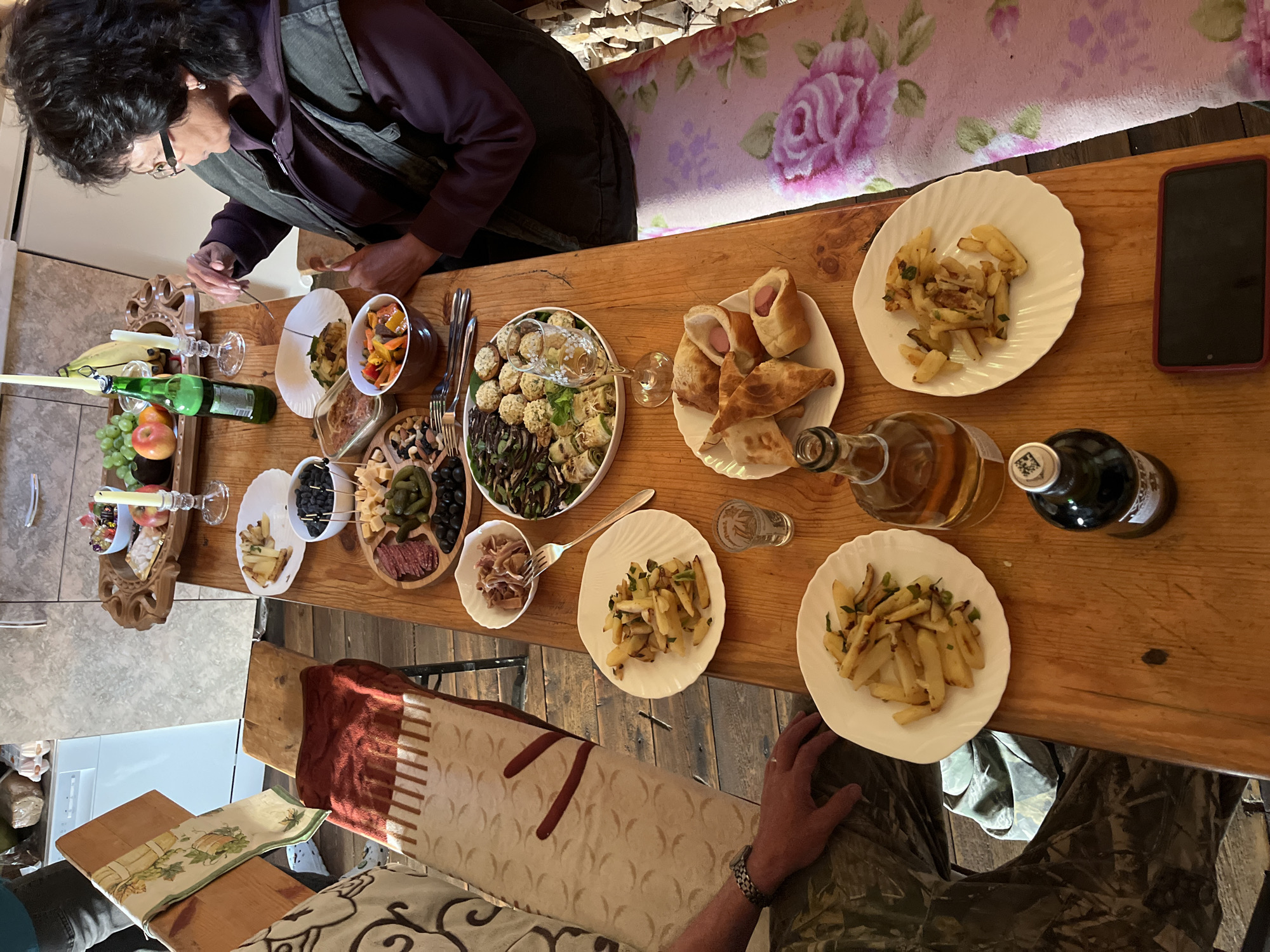 In Sovetskaya Gavan', Russia, a woman with dark, curly hair, wearing glasses and a dark purple hoodie with a vest, is seated at a wooden table laden with food. She is using a fork to pick up food from a white plate. The table is covered with various dishes, including plates of french fries, a charcuterie board with meats and cheeses, a bowl of olives, and a plate with pastries wrapped around hot dogs. There are also bottles of liquid, a wine glass, and a small glass of what appears to be vodka. In the background, a wooden bench is visible, draped with a patterned textile and a red blanket. The flooring is made of rough wooden planks. The scene is illuminated by natural light, creating a warm and inviting atmosphere.