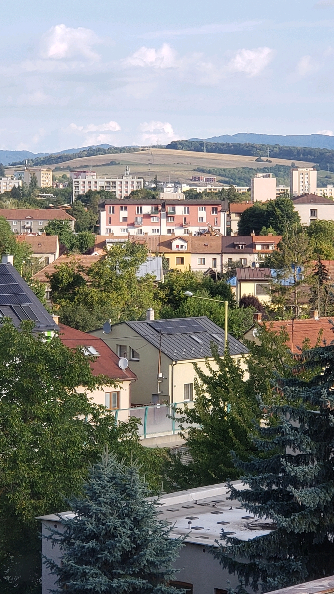 The image captures a panoramic view of a residential area in Prešov, Slovakia, with a mix of apartment buildings and houses nestled among lush green trees. In the foreground, the roofs of several houses are visible, some equipped with solar panels, suggesting a modern and eco-conscious approach to housing. The middle ground is dominated by a pink and white apartment building, characteristic of post-war architecture, surrounded by other residential structures and mature trees. The background reveals a rolling landscape of hills, partly covered by vegetation and featuring some distant buildings and infrastructure, possibly including communication towers or power lines. The sky is a bright blue with scattered white clouds, indicating fair weather. The overall scene depicts a well-established urban environment integrated with nature, presenting a calm and picturesque residential setting in Prešov.