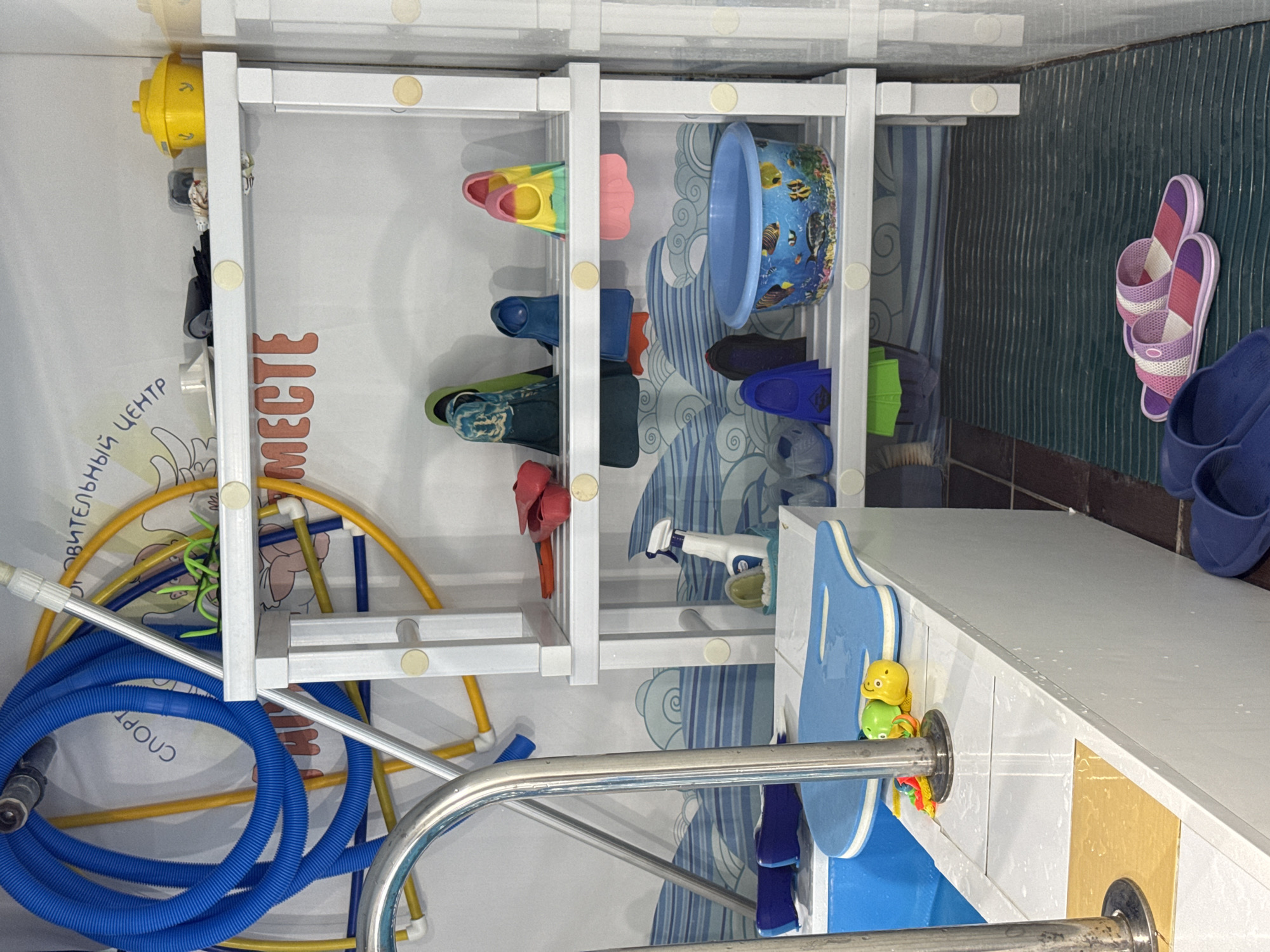 This image captures an organized storage area within what appears to be an indoor swimming facility, likely in Sochi, Russia, given the visible Cyrillic text. The scene is dominated by a white, multi-tiered shelving unit used to store various items related to swimming.
On the shelves, numerous pairs of swim fins (flippers) are neatly arranged, showcasing a variety of colors including yellow, pink, blue, green, red, and dark blue. A blue plastic bucket, decorated with an underwater scene featuring fish and coral, sits on one of the shelves. Other items include blue plastic clogs/sandals and a white spray bottle.
To the right of the shelving unit, against a dark, textured wall, two pairs of sandals are propped up: one pair with pink and purple stripes, and another dark blue pair.
To the left, coiled blue and yellow hoses, possibly for pool maintenance or cleaning, are visible alongside a silver metal pole. The wall behind these items features light-colored wallpaper with abstract blue wave patterns and cartoonish elements, including bananas and figures.
Along the bottom of the image, a white counter or ledge supports a blue kickboard or float. Two small, colorful rubber toys, a yellow and a green character, are stacked near a prominent metal handrail, typical of a pool deck.
Visible text on the wallpaper includes partially obscured Cyrillic words: "ЬНЫЙ ЦЕНТР" (likely part of "СПОРТИВНЫЙ ЦЕНТР" - Sports Center) and "МЕСТЕ" (likely part of "ВМЕСТЕ" - Together). The word "СПОРТ" (Sport) is also visible on the blue hose.
The overall impression is that of a functional and well-maintained area for storing swimming equipment, possibly for a children's swim school or a public pool. No people are present, and the artificial lighting suggests an indoor setting.