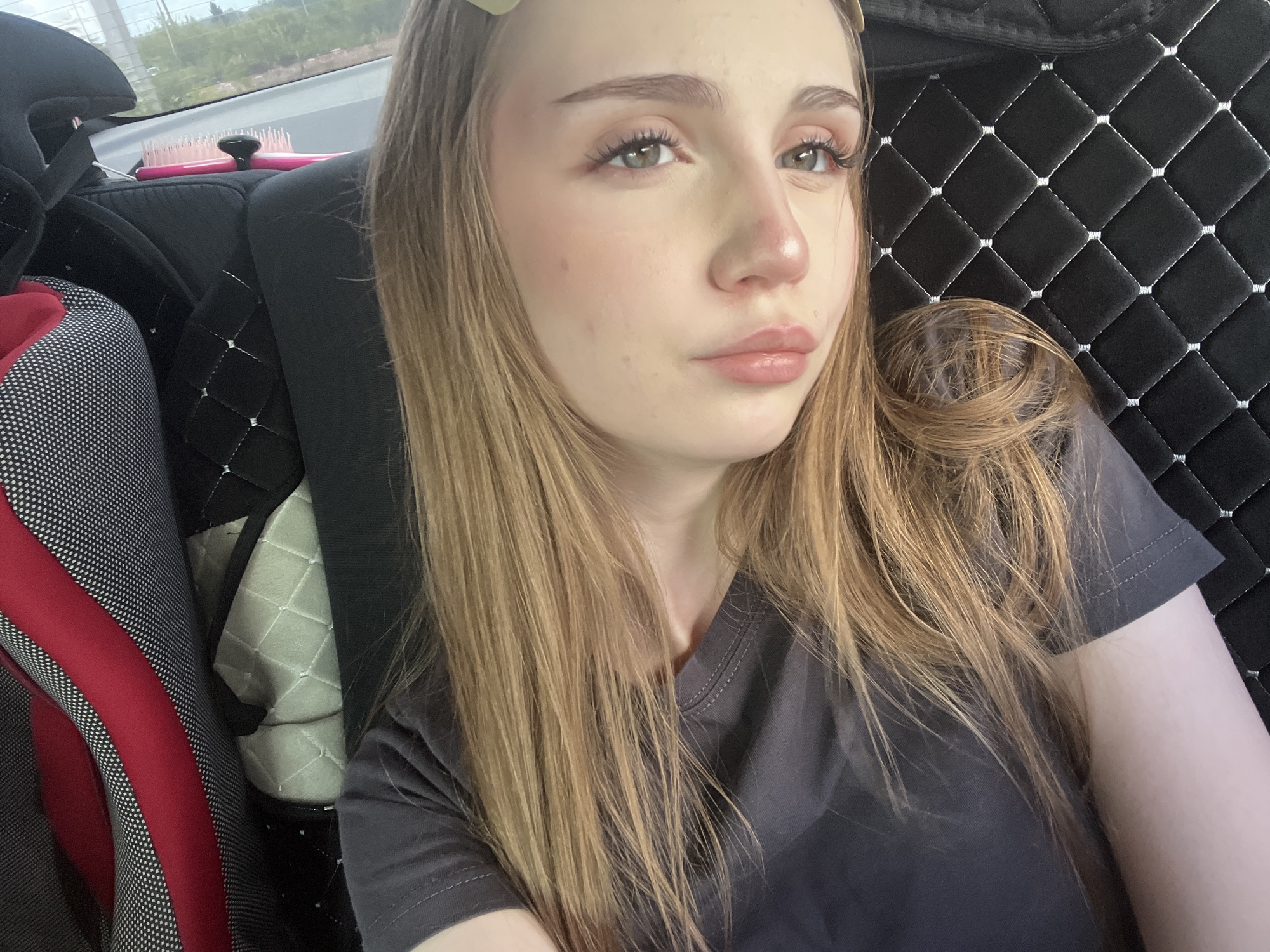 A young woman with long, blonde hair is seated in the backseat of a vehicle, looking towards the camera. She is wearing a dark gray t-shirt. The interior of the car features black quilted upholstery on the seat and a portion of a car seat with red and gray accents is visible. A pink hairbrush rests on the seat behind her. Outside the car window, trees and greenery are visible, suggesting a location with some natural surroundings, possibly on the outskirts of a populated area, such as near Новая Отрадовка, Russia. The lighting is natural, likely daylight.