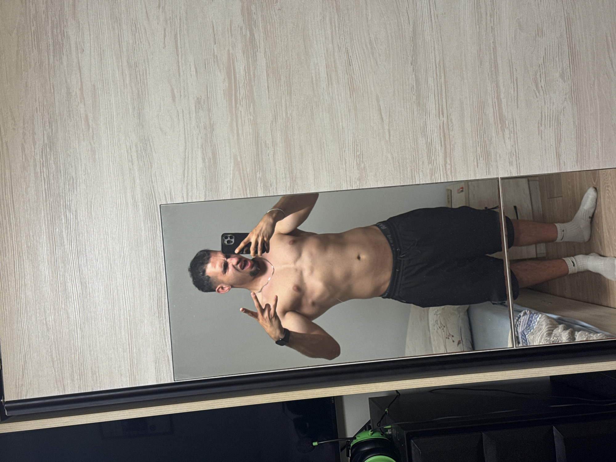 A shirtless man stands in front of a mirror, taking a selfie. He has short dark hair and a beard, and is wearing black shorts and white socks. He is making a hand gesture with his right hand. The mirror reflects a bedroom with a bed and a nightstand. The background is a wooden closet. The image is taken from a medium shot. The location is not specified in the image or surrounding context.