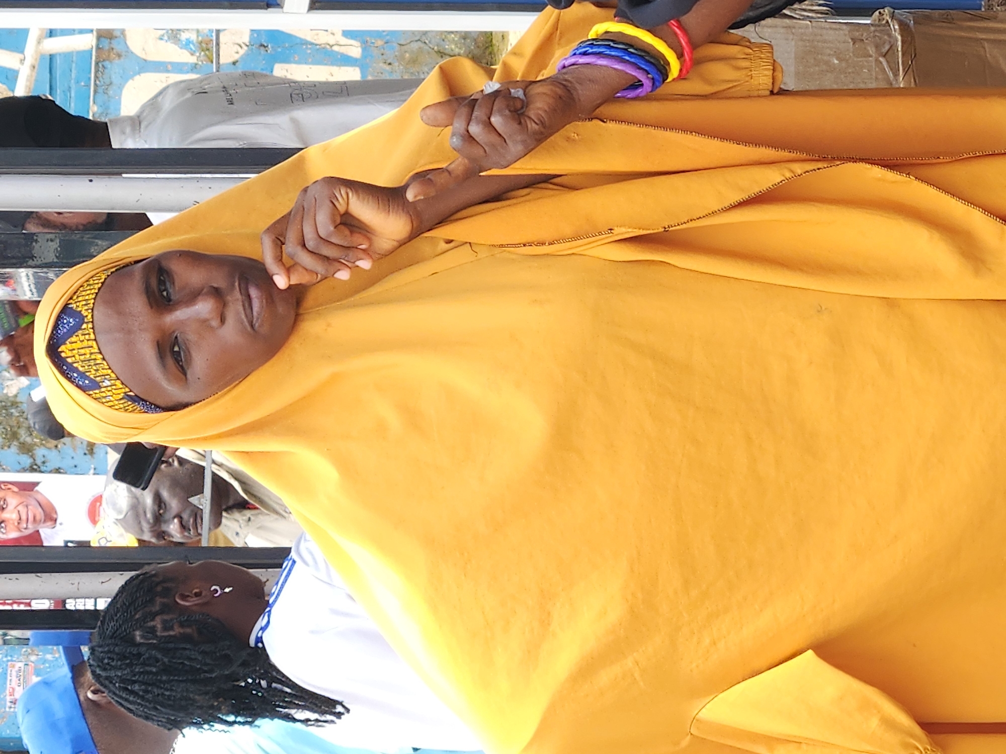 In Jos, Nigeria, a woman wearing a yellow hijab and an orange dress stands with her right hand raised, fingers extended, as if making a point or giving a gesture. She has a thoughtful expression on her face, with her chin resting on her hand. The background is blurred, but suggests an outdoor urban setting with other people visible, including a person in a blue cap and a man with a black hat, possibly in a market or public gathering area. The woman is adorned with colorful bracelets on her right wrist: purple, blue, yellow, and red.