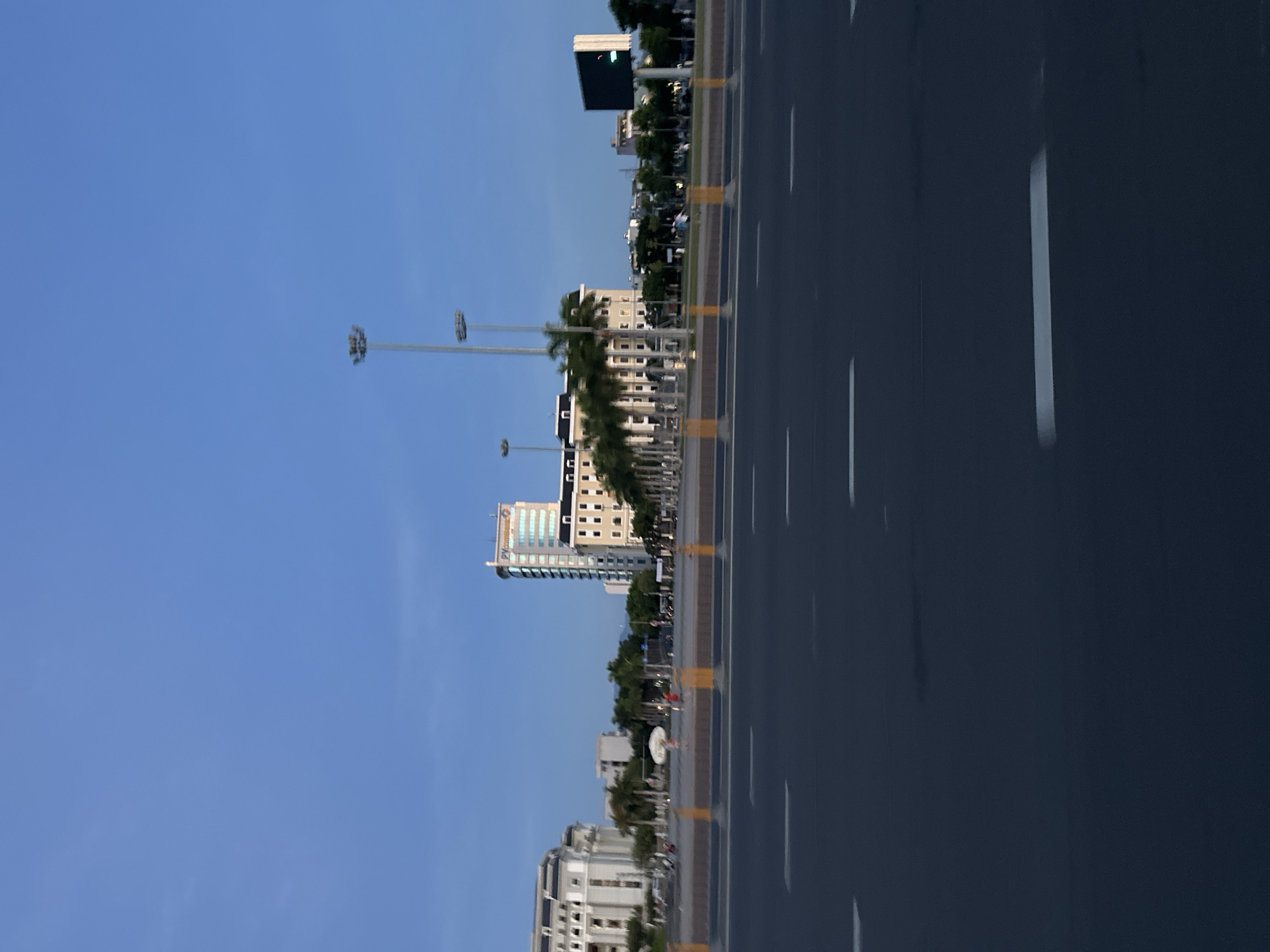 The image depicts a wide, dark asphalt road with white lane markings, suggesting a city street or highway in Đà Nẵng, Vietnam. The perspective is from a vehicle in motion, indicated by the slight blur in the foreground and the sweep of the road. Across the road, a pedestrian area or plaza is visible, separated by a median with yellow bollards.
Beyond the plaza, several buildings form the city skyline. On the left, a grand, white building with multiple stories and arched windows stands prominently. To its right, a row of palm trees lines the plaza, leading towards a lighter-colored, mid-rise building with a grid of windows. Further to the right, a taller, modern skyscraper with a reflective facade and a visible "PVcombank" logo dominates the scene. Adjacent to it is another cream-colored building with a tiled roof. A large digital billboard with a green light is positioned to the far right.
In the distance, a faint mountain range is visible under a clear blue sky. Two tall, modern lampposts with multiple light fixtures stand in the middle ground. Several people are visible walking in the plaza area, and a few vehicles are parked or driving in the distance, though they are indistinct due to the distance and motion blur. The overall atmosphere suggests an urban environment in the late afternoon or early evening, with the sky transitioning from day to dusk.
