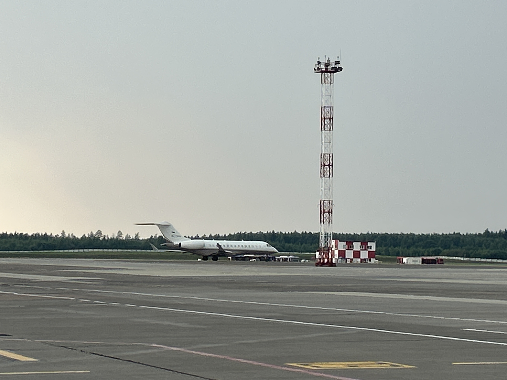 This is a daytime, eye-level, medium-shot photograph taken from an airport tarmac. In the mid-ground, a white private jet with its tail visible is parked on the tarmac. The jet has dark windows and appears to be a business jet. To the right of the jet, a tall, red and white checkered communication tower stands prominently. Further in the background, a line of green trees forms a backdrop against a pale, overcast sky. The foreground consists of the asphalt surface of the airport runway, with visible white and yellow markings. The scene is devoid of people. The image appears to have been captured at an airport, likely Minsk National Airport (MSQ) in Minsk, Belarus, given the presence of airport infrastructure and typical foliage for the region.