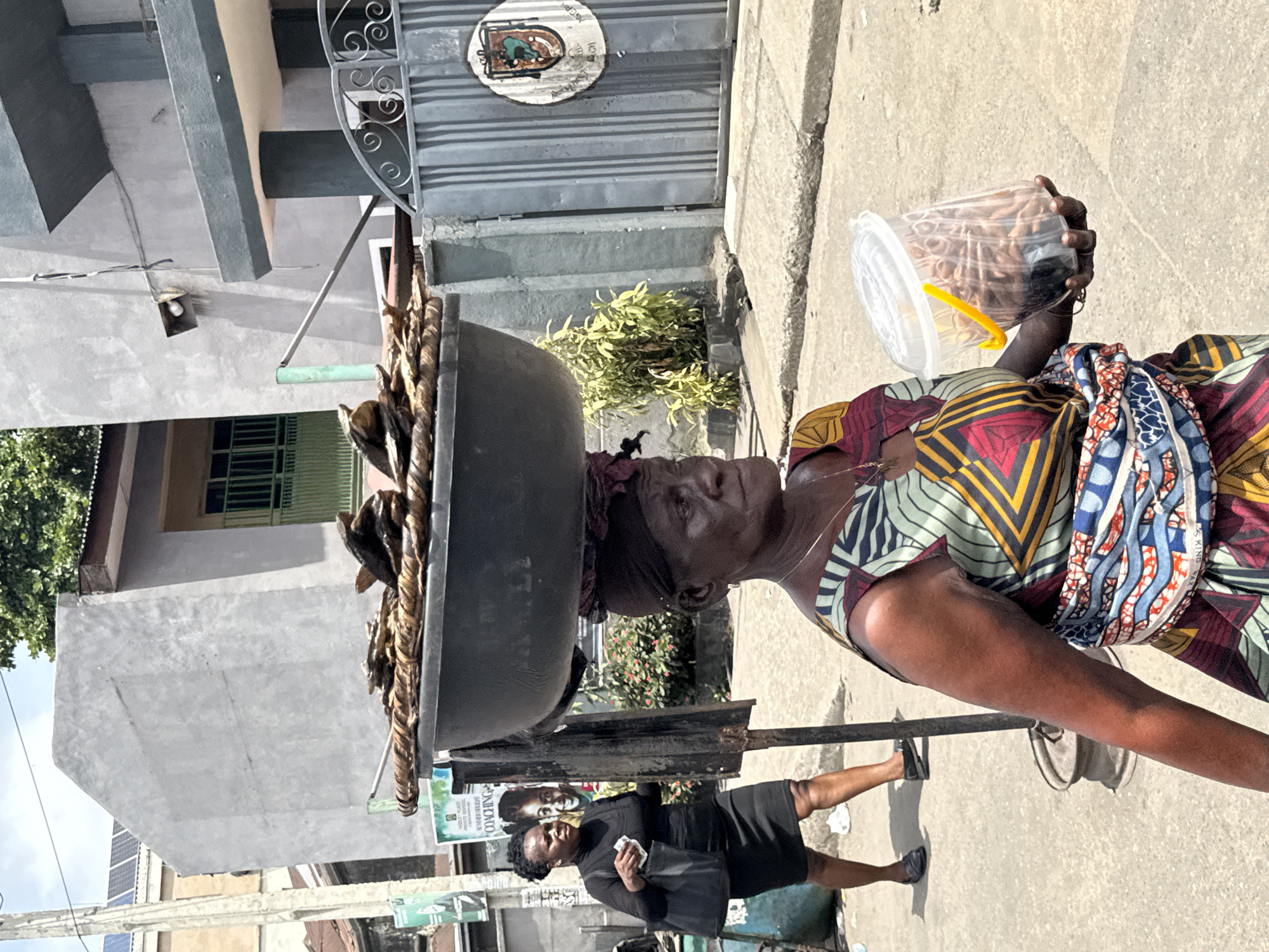An elderly African woman in Lagos, Nigeria, carries a large bowl atop her head, likely filled with goods for sale, and a clear plastic container filled with snacks in her hand. She wears a colorful patterned dress and a headwrap. A younger woman in a black dress walks nearby. The setting is a city street with buildings, a gate, and advertisements visible.