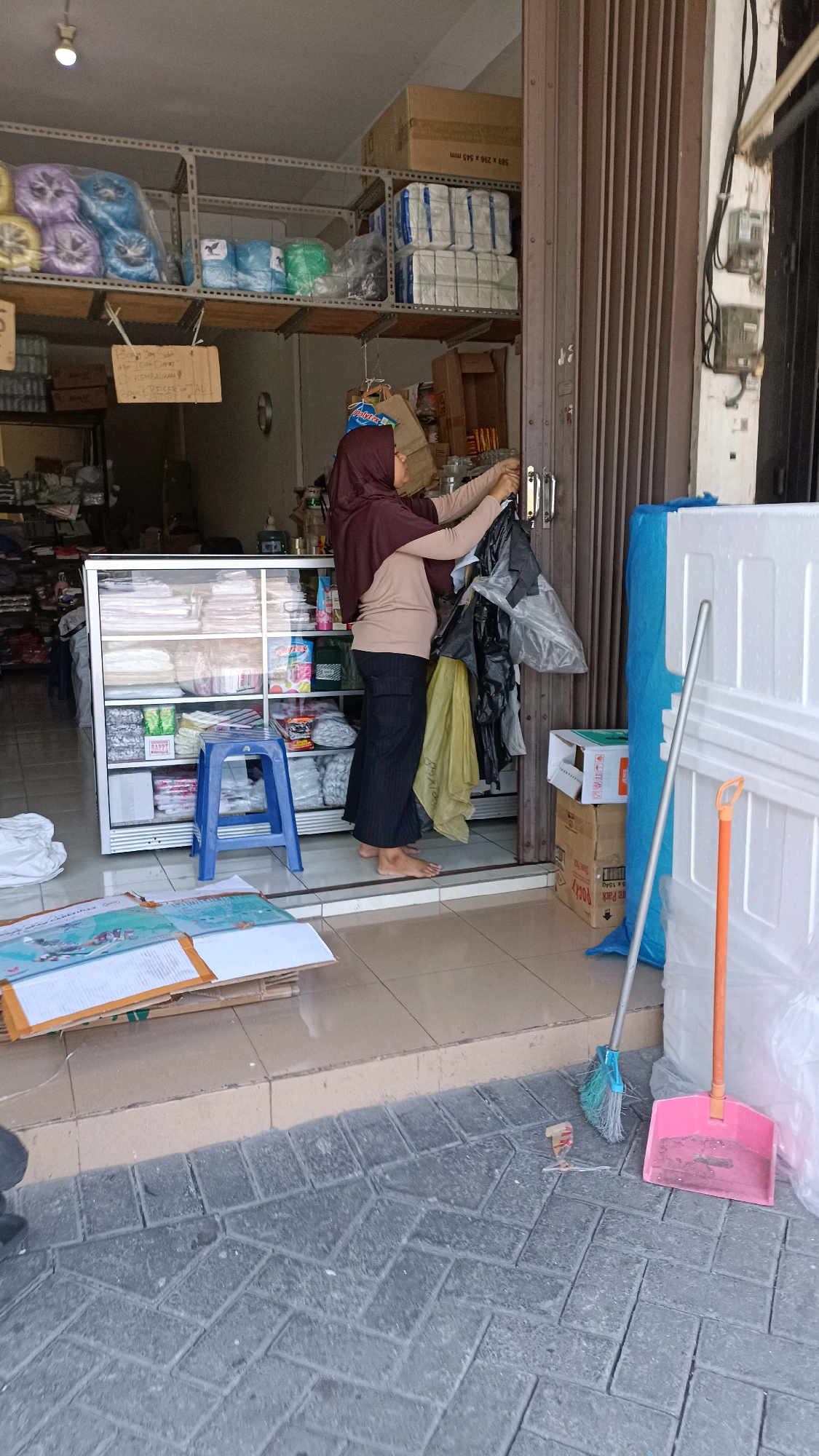 The image depicts the exterior of a small retail establishment, likely a shop in Surabaya, Indonesia, situated at ground level with a rolling metal door partially open. A woman wearing a brown hijab and a light brown top paired with dark pants is seen standing at the entrance. She is holding several black plastic bags, appearing to be either putting them away or taking them out. In front of the shop, a display cabinet filled with various packaged goods is visible. A small blue plastic stool is placed in front of the cabinet. The shop's interior, visible through the open doorway, contains shelves stocked with items, including rolls of colorful material on an upper shelf and boxes stacked on the floor. Outside the shop, on the paved walkway, a broom with a blue handle and green bristles and a pink dustpan are present, suggesting recent cleaning or preparation for cleaning. Cardboard boxes and large white styrofoam packaging are stacked to the right of the entrance. The overall scene suggests a commercial area with typical retail operations.