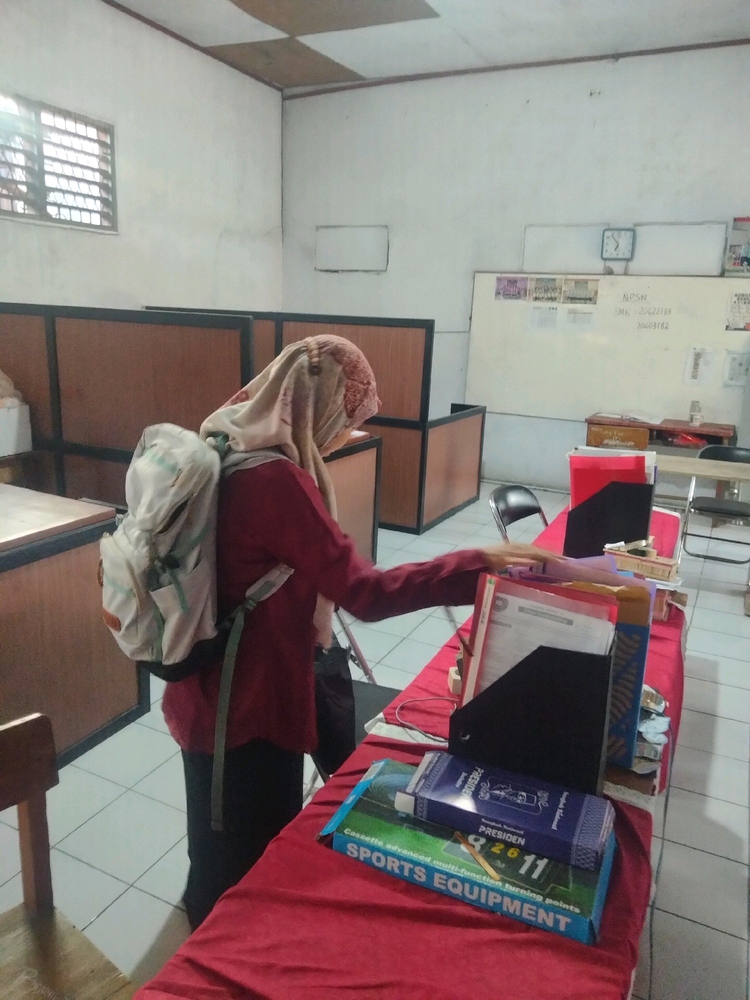 The image depicts an indoor scene, possibly a classroom or office, in Suka Sari, Indonesia. A person wearing a maroon shirt and a patterned headscarf is standing and looking towards a table covered with a red cloth. The person is carrying a light-colored backpack. On the table, there are several folders, books, and other office supplies. One of the books has the text "PRESIDEN" and "SPORTS EQUIPMENT" visible on its cover. In the background, there is a window with a grille, a whiteboard with some writing and a clock, and wooden partitions. The floor is tiled. The lighting suggests it might be daytime, with light coming from the window.