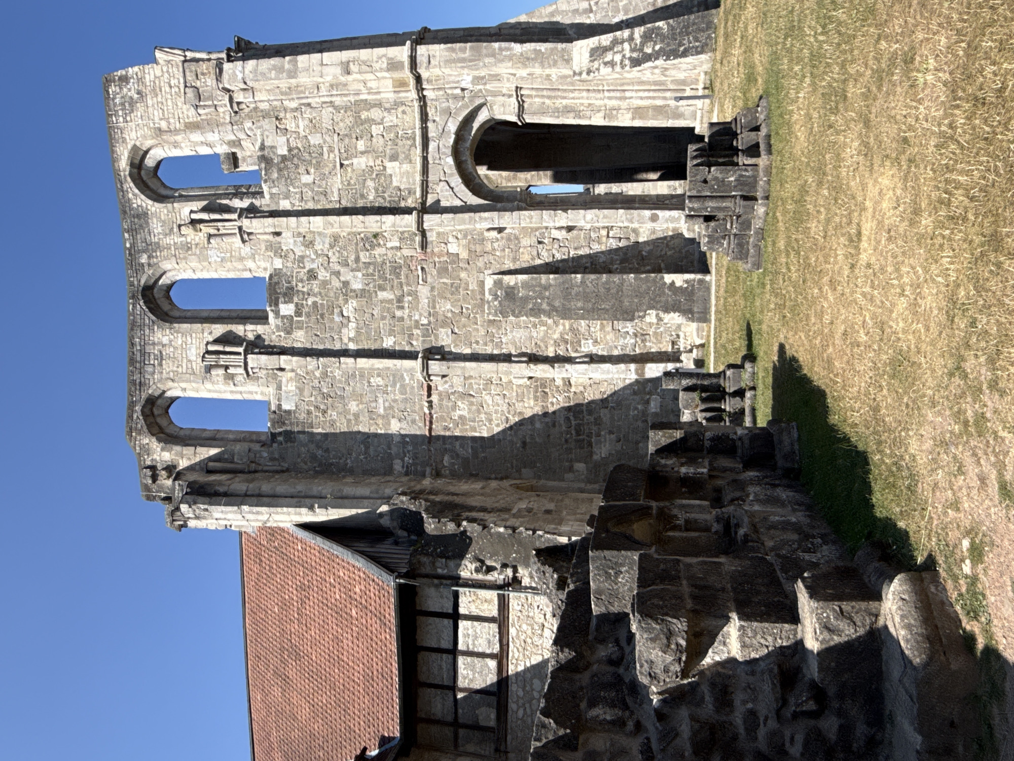 The image shows the exterior of the Walkenried Abbey ruins in Walkenried, Germany. The ruins are made of stone and feature arched windows and doorways. A partially preserved building with a red tiled roof and timber framing is attached to the ruins on the left. The ground in front of the ruins is covered in dry grass, and there are several stone fragments scattered across the area. The sky is clear and blue.