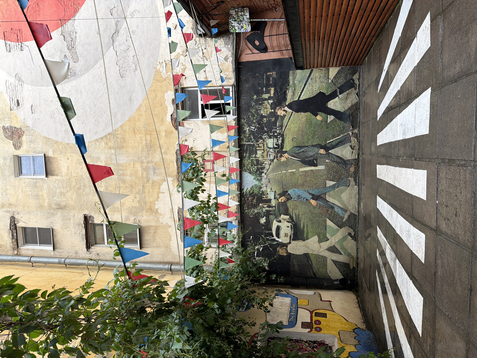 In a cobblestone courtyard in an unknown city, a mural depicting the iconic Abbey Road album cover of The Beatles adorns a weathered wall. The painting shows John Lennon, Paul McCartney, George Harrison, and Ringo Starr walking across a street. White stripes painted on the ground mimic a crosswalk, extending from the foreground towards the mural. Above the mural, a string of colorful triangular flags hangs, fluttering against the backdrop of aged, peeling walls and a few windows. Lush green foliage cascades from the left side of the image, partially obscuring a yellow and blue painted section of the wall that includes the words "The Beatles". The scene appears wet, suggesting recent rain, with reflections visible on the ground. To the right, a wooden fence with horizontal slats provides a contrast to the rougher textures of the building.