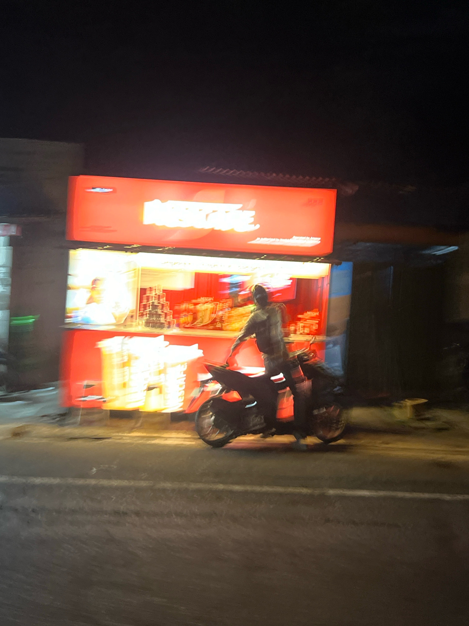 A nighttime street scene in Magelang, Indonesia, shows a person on a motorcycle passing a brightly lit food stall. The stall has a large red sign with white lettering, partially obscured by motion blur. Inside the stall, there are displays of products, possibly food items, in yellow and red packaging. A person is visible behind the counter, appearing to be a vendor. The motorcycle rider, also blurred due to motion, is wearing a light-colored shirt. The street is wet, reflecting the lights from the stall and the sky above is dark.