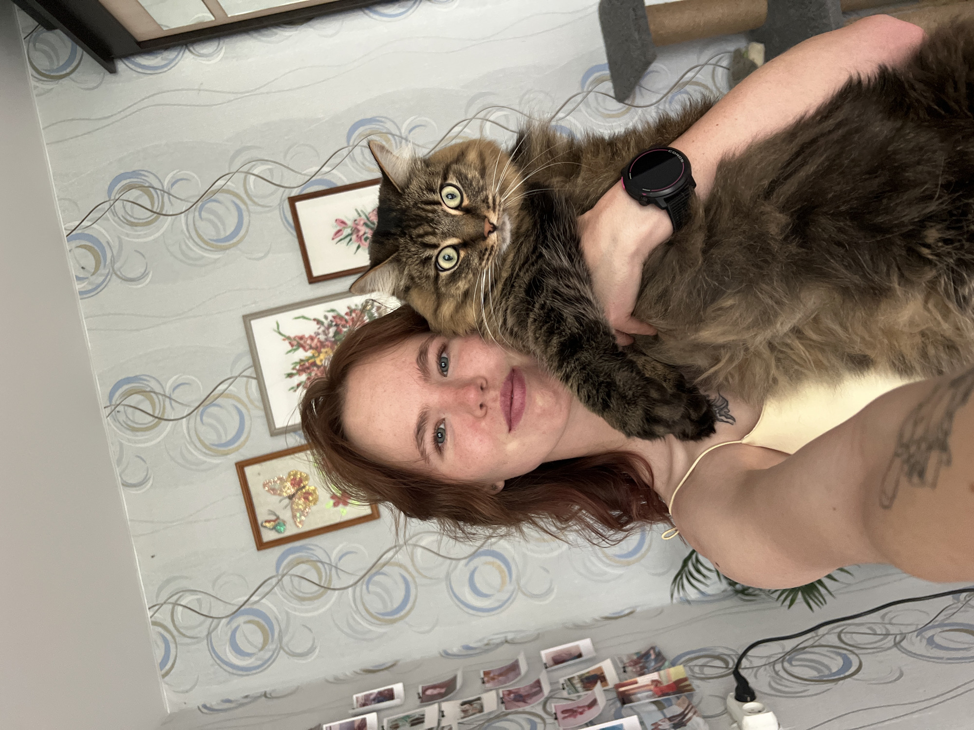 A young woman is holding a large, long-haired tabby cat in what appears to be an indoor setting in Chaikovsky, Russia. The woman, with reddish-brown hair, fair skin, freckles, and blue eyes, looks towards the camera with a neutral expression. She wears a light-colored top and has tattoos visible on her right arm and shoulder. On her left wrist, she wears a black smartwatch with a pink accent, with "COROS" faintly visible on its face.
The fluffy tabby cat, with prominent whiskers and bright green eyes, is held close to the woman's face and chest, staring directly at the camera with wide, alert eyes.
The background features light grey wallpaper patterned with swirling circles in light blue, beige/gold, and grey, interspersed with wavy lines. Several framed pictures of floral and butterfly designs are hung on the wall. A black electrical cord runs across the wallpaper, leading down towards a partially visible outlet. Part of a cat scratching post or tree is visible in the upper right. Below the subjects, a surface displays a collection of small printed photos, possibly strung along lines. The image captures a casual, intimate moment between a pet owner and their cat.