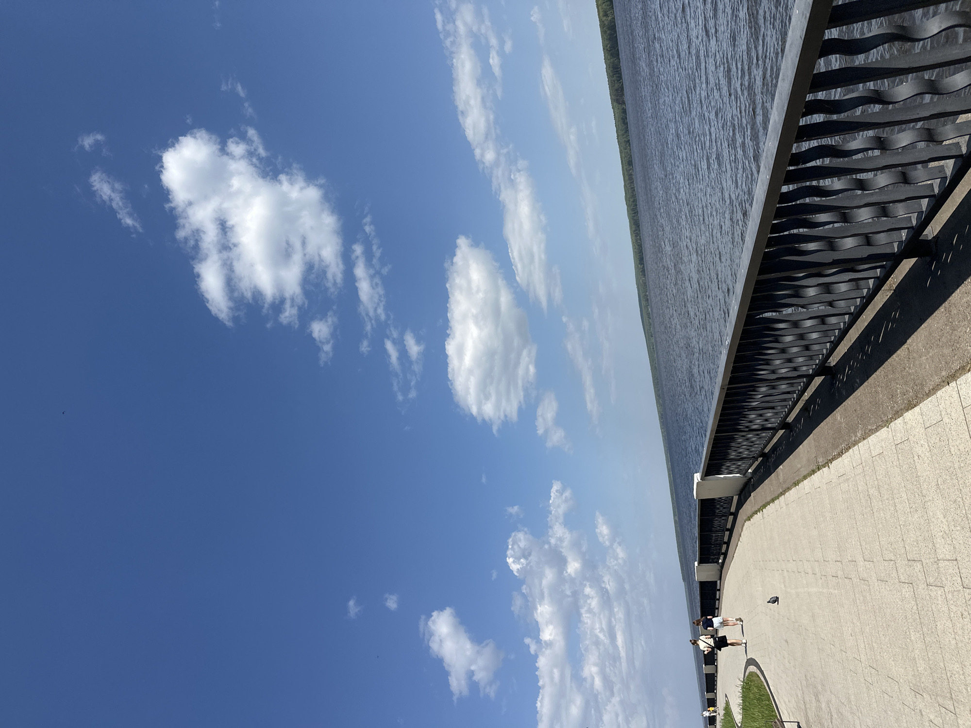 The image depicts a waterside promenade in the city of Chaikovsky, Russia, during daylight hours. The view includes a section of the city's embankment with a modern black metal railing, a paved walkway, and a body of water adjacent to the embankment. The sky is predominantly blue with scattered cumulus clouds.
Two people are walking along the walkway, and a pigeon is also present. The walkway appears to be constructed of light-colored paving stones. Some greenery is visible between the walkway and seating areas. The water is dark and calm. The edge of the opposite shore is lined with trees.