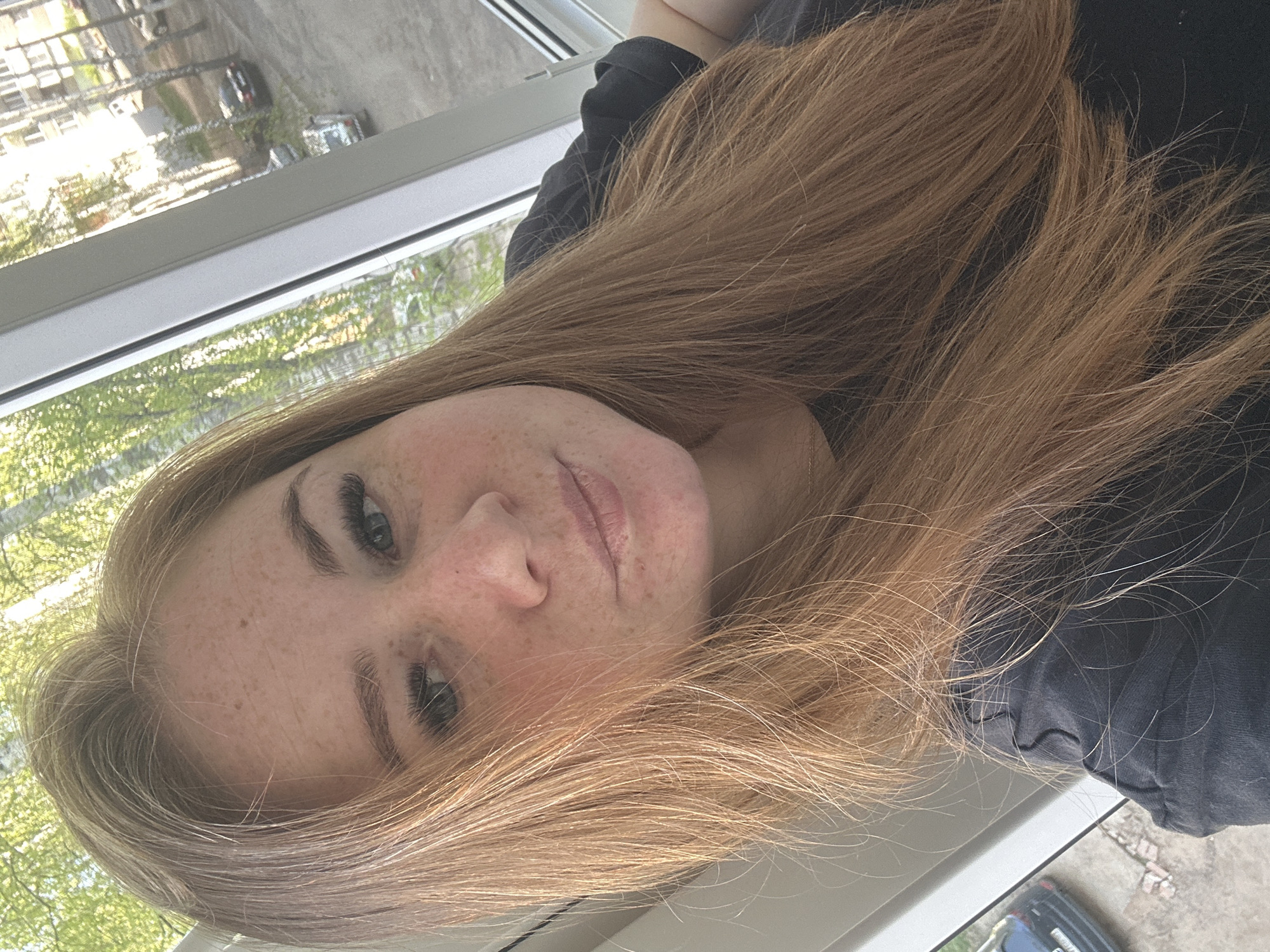 A woman with freckled skin and light brown hair is indoors, presumably in an apartment. She is wearing a black shirt. A window is visible in the background, offering a view of trees and buildings. Based on the information provided, it is not possible to determine the exact location in Chaikovsky, Russia without further information.