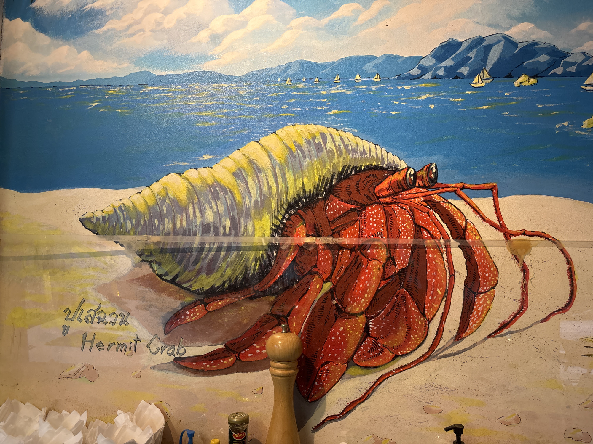 A mural on a wall in Chalong, Thailand, depicts a vibrant beach scene. The focal point is a large, bright red hermit crab with white speckles, positioned on a sandy shore. Its shell is a prominent, curled structure with yellow and grey stripes. In the background, a calm blue ocean stretches towards distant, hazy mountains. Several white sailboats are dotted across the water. Above the ocean, white clouds drift in a pale blue sky. Text written on the wall beneath the crab includes Thai script and the English words "Hermit Crab." The foreground shows part of a wooden pepper grinder and a small metal spice container.