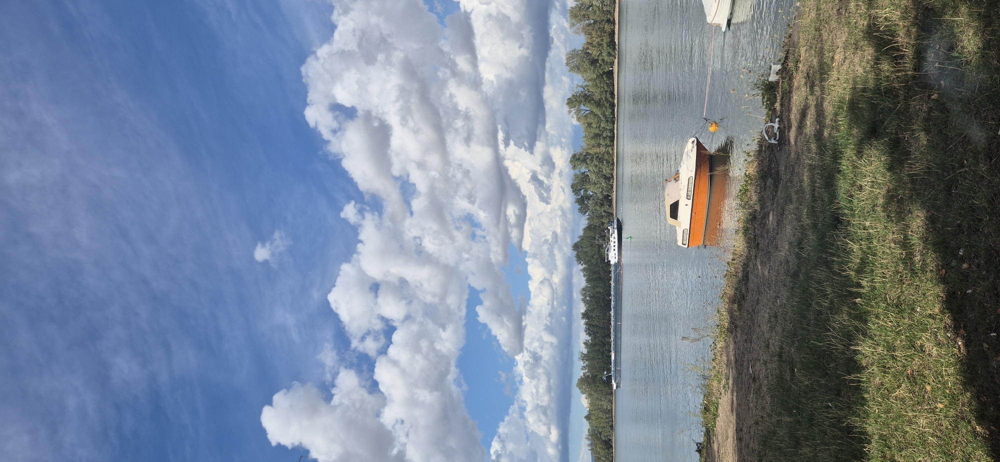 The image captures a serene daytime scene on a river, likely the Rhine in Mainz, Germany, judging by the presence of a barge and a pleasure craft typical of German waterways. The sky is a vibrant blue, adorned with dramatic cumulus clouds that cast shadows on the water. In the distance, a long, dark barge navigates the river, followed by a smaller tugboat. Closer to the foreground, an orange and white motorboat is moored, its reflection shimmering on the calm surface of the water. The riverbank is lined with lush green grass, with trees forming a dense backdrop along the opposite bank. A few discarded ropes and white objects are visible on the grass near the water's edge. The overall atmosphere is peaceful and tranquil, suggesting a pleasant day for boating.