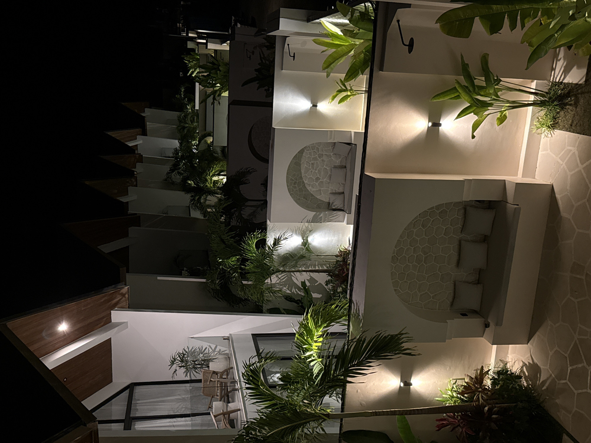 This media file captures a modern resort or hotel complex at night, located in Kuta Selatan, Indonesia. The scene is illuminated by warm, artificial lighting, highlighting architectural details and tropical vegetation against a dark sky.
The multi-story building features a contemporary design with white walls, clean lines, and some dark and wooden accents. Each unit appears to have an outdoor seating area or balcony, equipped with light-colored cushions and possibly woven furniture, visible in various crops. A distinctive design element is the circular, textured wall art or pattern, resembling a mosaic or stone arrangement, often placed behind these seating areas.
Lush tropical plants, including palm trees, broad-leafed foliage, and smaller shrubs, are integrated throughout the complex. These plants are strategically lit by upward and downward-facing wall sconces and recessed lighting, casting dramatic shadows and creating depth. Glass railings are visible on some balconies, offering an open feel.
No people or specific activities are depicted; the scene emphasizes the architectural and natural beauty of the resort environment. The lighting creates a serene and inviting atmosphere, typical of a luxury vacation destination. No text is visible in the image.