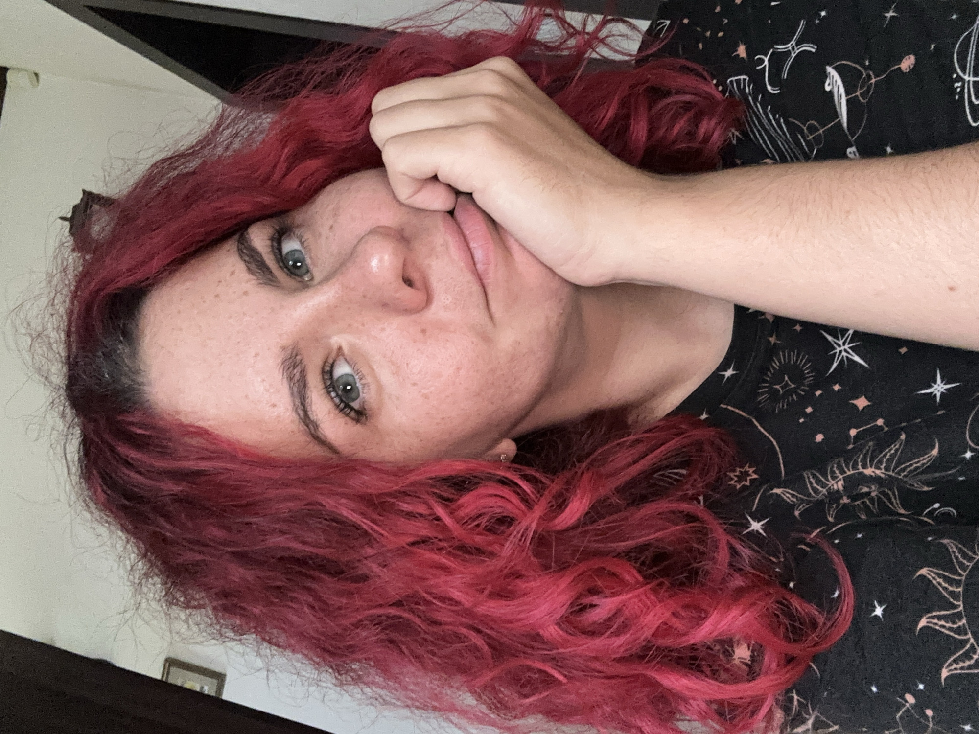 A close-up portrait of a woman with red, curly hair resting her chin on her hand. Her eyes are light-colored, and she has freckles on her face. She is wearing a black shirt with celestial designs. The background shows a wall and a dark-colored object, possibly furniture. The location cannot be determined.