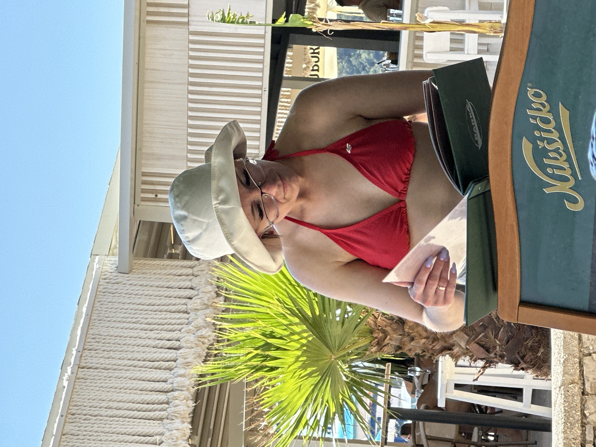 In Boreti, Montenegro, a woman wearing a red bikini and a white sun hat is looking down at a green menu. She has on glasses and her nails are painted lavender. A gold ring is on her left ring finger. She is holding the menu open with her left hand. The menu is on a table, and a wooden frame is visible at the bottom of the image. Behind her, a palm tree with green fronds is illuminated by the sun, and a light-colored building with vertical striped paneling and a decorative rope fringe is visible. The sky is clear and blue.