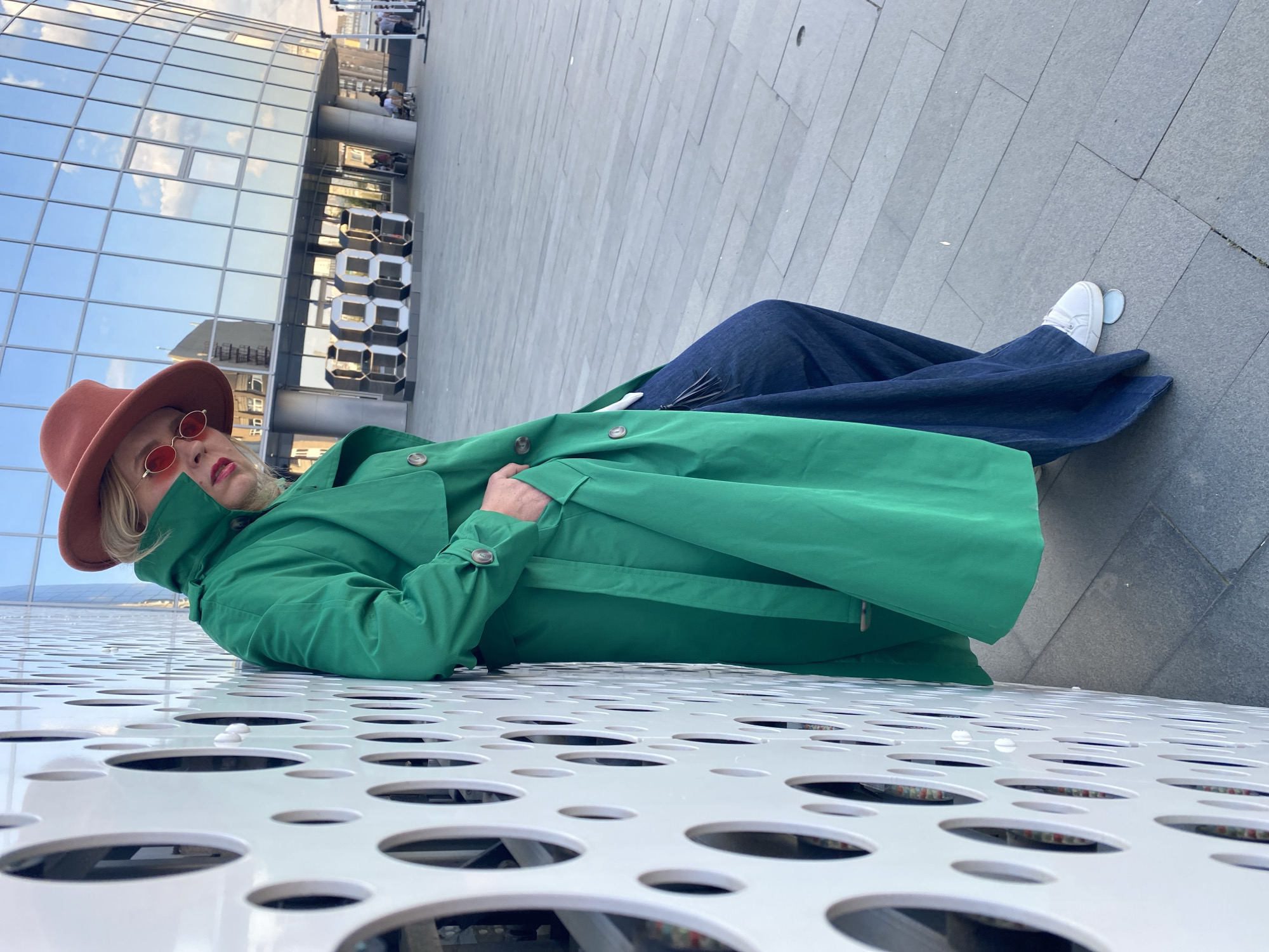 The image captures a person lying on a white, perforated metal surface outdoors. The individual is wearing a green trench coat, a brown hat, and round, red sunglasses. They also appear to be wearing wide-leg denim pants and white sneakers.
In the background, there's a modern glass building with a distinctive silver digit sculpture representing "8888" next to it. The location is the Embankment of the Iset River in Yekaterinburg, Russia, close to the Presidential Center of Boris Yeltsin. The photograph's perspective is unusual, as if it were taken from a low angle, emphasizing the patterned metal surface and the person lying on it.