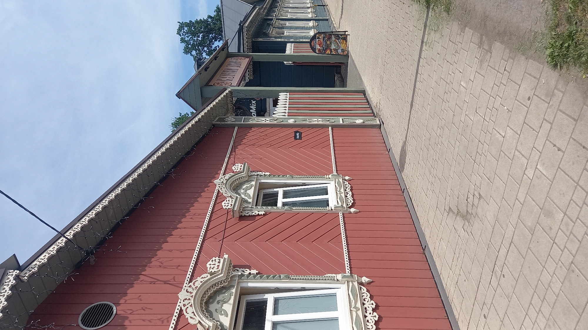 This image depicts a street scene in Zvenigorod, Russia, featuring two historic wooden buildings with intricate architectural details under a clear sky.
The prominent structure on the left is a single-story wooden building painted a deep red or maroon. Its exterior walls feature distinct siding patterns: the upper section consists of diagonal planks, while the lower section is composed of horizontal planks. Two white-framed windows are visible on this wall, each adorned with elaborate, light-colored carved wooden platbands that extend above and below the window opening. The left window is slightly ajar, opening inward, while the right window is also open, tilted from the top. A small, white, rectangular sign with the text "AsiaMix" in black is affixed to the wall between these two windows. Near the upper left corner of the building, a circular, louvered metal vent is installed. Along the eaves, decorative white carved trim in a scalloped pattern is visible, beneath which a string of small, white festive lights is strung. A black utility wire extends diagonally from the roofline towards the upper left sky.
To the right of the red building, an entrance or gate structure leads to another building. This entrance features a gabled roof in green and dark teal, with a sign mounted beneath the gable that reads "КУЛИНАРНЫЙ ДОМ" (Kulinarny Dom), translating to "Culinary House." The gateposts and sections of the fence leading to this entrance are painted with vertical stripes of green, red, and white. Beyond this gate, a dark teal or blue wooden building is visible, also featuring multiple white-framed windows with carved surrounds, similar in style to those on the red building. A freestanding A-frame sign for "AsiaMix," displaying food images, stands on the sidewalk near the entrance.
The ground in the foreground is a paved sidewalk constructed from light-colored interlocking pavers of various rectangular and square shapes, exhibiting some minor wear and discoloration. A small patch of green grass and weeds protrudes from a crack in the pavement in the mid-ground. Along the far right edge of the image, a strip of green grass is visible. The sky above is a bright, pale blue with scattered wisps of white clouds. In the background, above the blue building, dark green deciduous trees are present. No people or vehicles are visible in the scene.