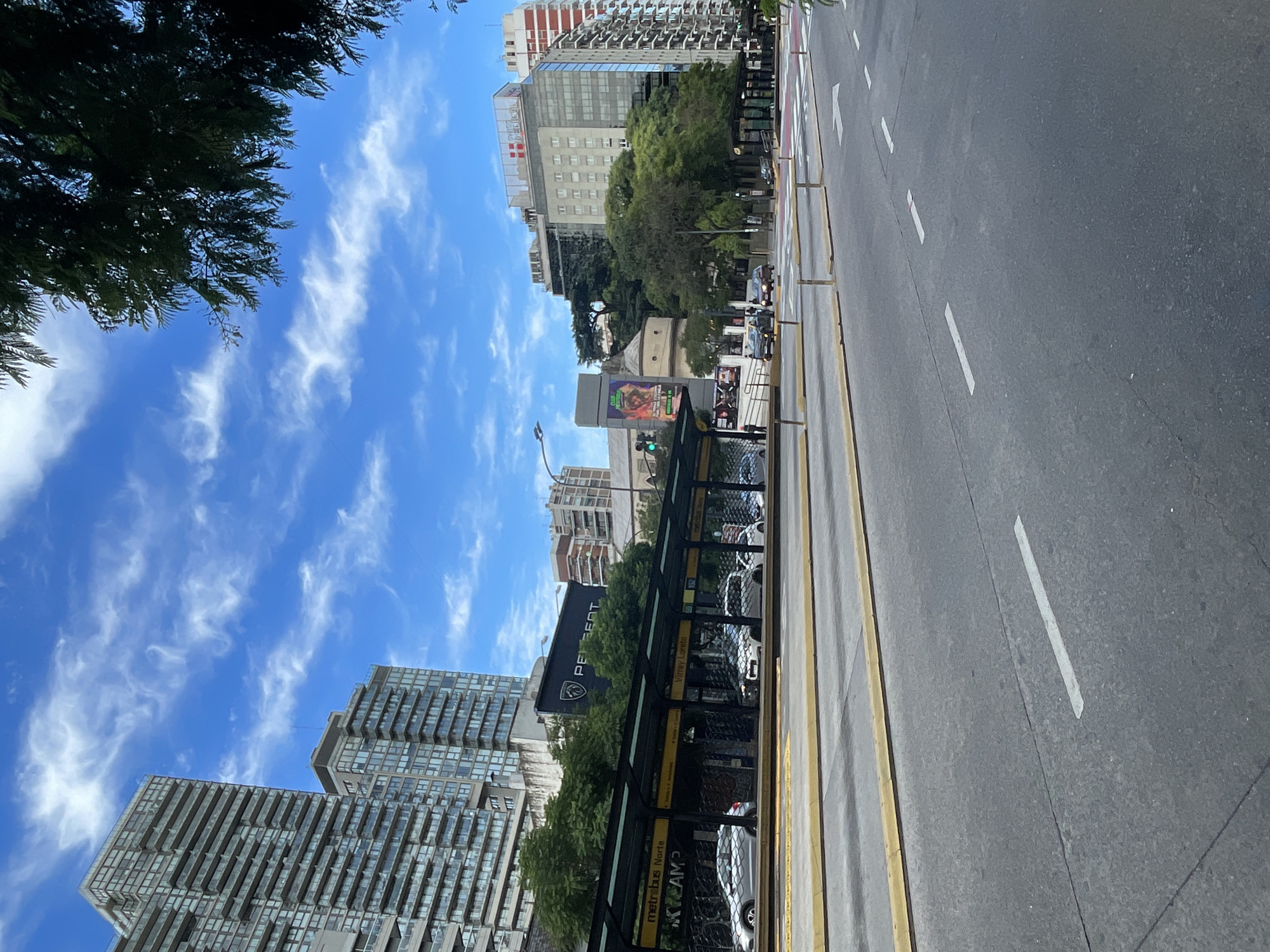 This street-level image shows a street scene in Buenos Aires, Argentina. A wide asphalt road with lane markings stretches to the right, bordered by a yellow median. On the left, a bus stop with a "metrobus Norte" sign and a Peugeot logo shields waiting passengers. Buildings of varying heights line the background, including residential apartments and commercial structures. The sky is bright blue with scattered white clouds. Trees are visible along the roadside, adding a touch of green to the urban environment. People and vehicles are present in the distance, suggesting an active city environment.