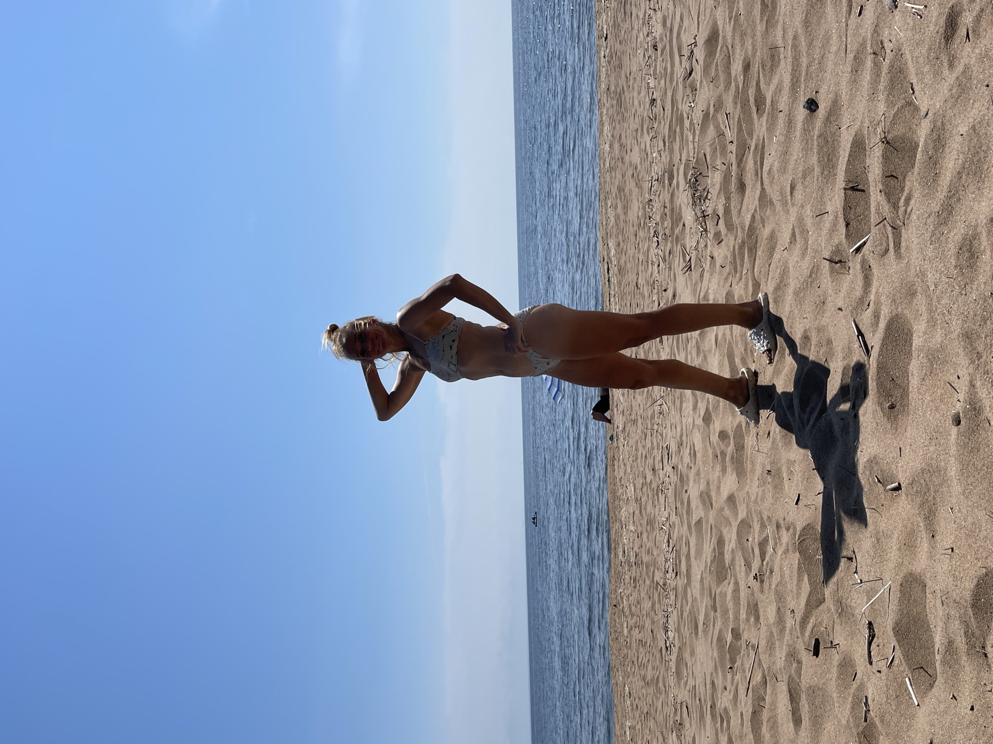 A woman stands on a sandy beach, posing with her hand on her hip and the other touching her head. She is wearing a light-colored bikini. The beach has scattered debris and footprints on the sand. In the background, the ocean stretches to the horizon under a clear blue sky. There are a few other people visible in the distance near the water, and an umbrella on the sand. The scene appears to be a sunny day at the beach in Zelenaya Roshcha, Russia.