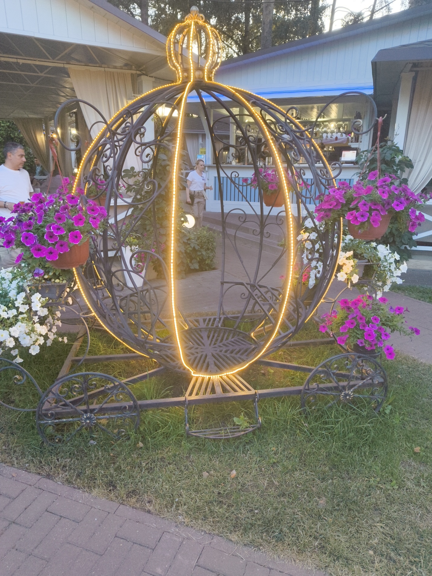 A decorative, illuminated carriage-shaped archway stands on a grassy area adjacent to a paved walkway. The archway is made of dark metal with intricate scrollwork and is adorned with a string of yellow lights that form a crown at the top. Potted plants with vibrant purple and white flowers are hung from the sides of the archway. In the background, a white building with a blue-lit awning and a serving counter is visible, suggesting it is an outdoor cafe or food establishment. Two individuals are present: one man in a white shirt is partially visible on the left, and a woman in a white top and khaki pants is seen walking in the background towards the right. The scene is set outdoors, likely in the late afternoon or early evening, with trees visible in the distance. This decorative element appears to be located in the city of Ramenskoye, Russia, as indicated by the prompt.