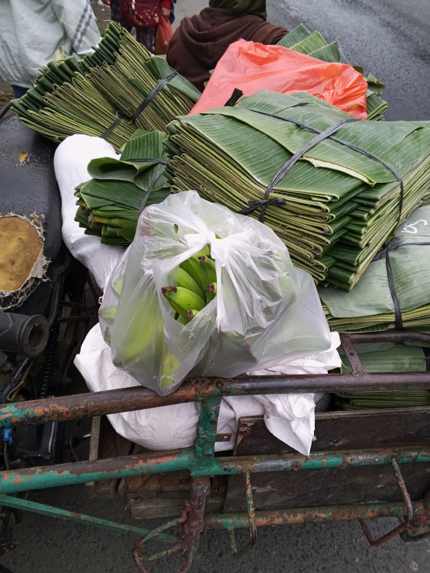 In Pancur Batu, Indonesia, a close-up shot captures a side view of a motorized cart laden with goods. A bundle of folded green banana leaves, secured with black twine, is prominently displayed. Adjacent to this, a clear plastic bag contains a bunch of green bananas. More stacks of folded banana leaves, some tied with black twine and one covered with an orange plastic sheet, are visible. The cart's rusty metal frame, painted green in some areas, supports the cargo. Partially visible in the background, a person in a brown garment and another in a white and striped garment suggest a market or roadside setting. The ground appears to be asphalt.