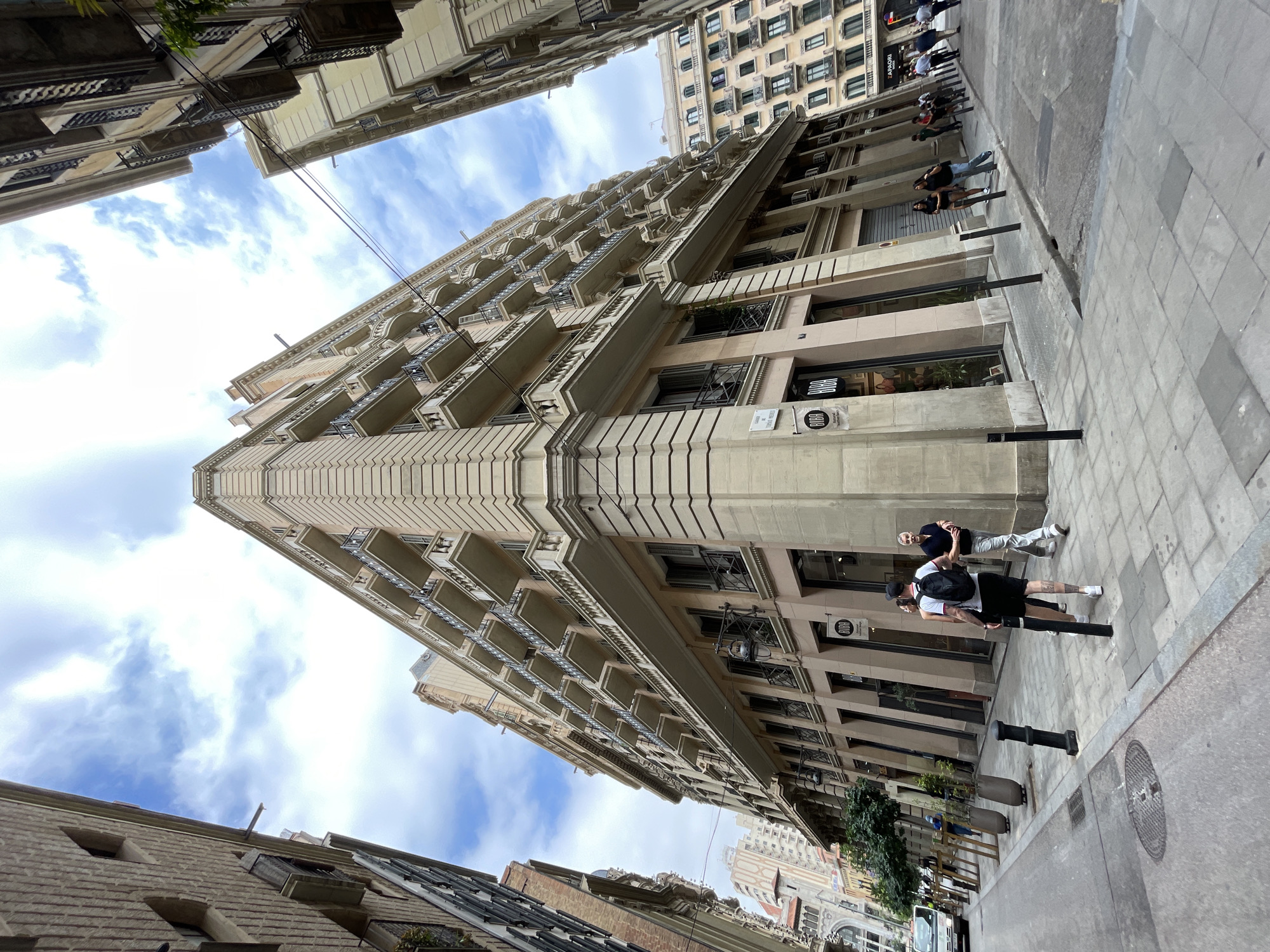 This media file captures a street scene in Barcelona, Spain, with a strong emphasis on the architecture of the buildings. The perspective is from a low angle, looking up towards the sky and along a street lined with tall, ornate buildings. The sky is partly cloudy, suggesting it's daytime, likely late morning or afternoon, with good natural light.
The main architectural feature is a grand, multi-story building with numerous balconies featuring decorative railings and cornices. The building's facade is a light beige or cream color, and it exhibits classical architectural elements. In the background, another building with a grid-like pattern of windows is visible, adding to the urban density of the setting.
Several people are present on the sidewalk. Some are walking, while others are standing, seemingly engaged in conversation or taking a break. Two individuals in the foreground are particularly noticeable: one wearing a black t-shirt, black shorts, a black cap, and a backpack, and another wearing a dark top and light-colored pants. They appear to be walking or standing casually. Further down the street, more people can be seen, some in groups, indicating a normal day with pedestrian activity.
Notable details include several black bollards lining the sidewalk, a manhole cover, and what appear to be air conditioning units attached to the buildings. There is also some greenery, possibly trees or shrubs, visible in the lower parts of the frame.
There is some visible text on a sign attached to the beige building, which appears to read "BJORG." Another, smaller sign next to it has a circular logo with text inside. These suggest commercial establishments on the ground floor of the buildings.
The overall impression is of a bustling urban environment in Barcelona, characterized by its distinctive architecture and everyday street life. The scene is vibrant and active, without any specific event being depicted.