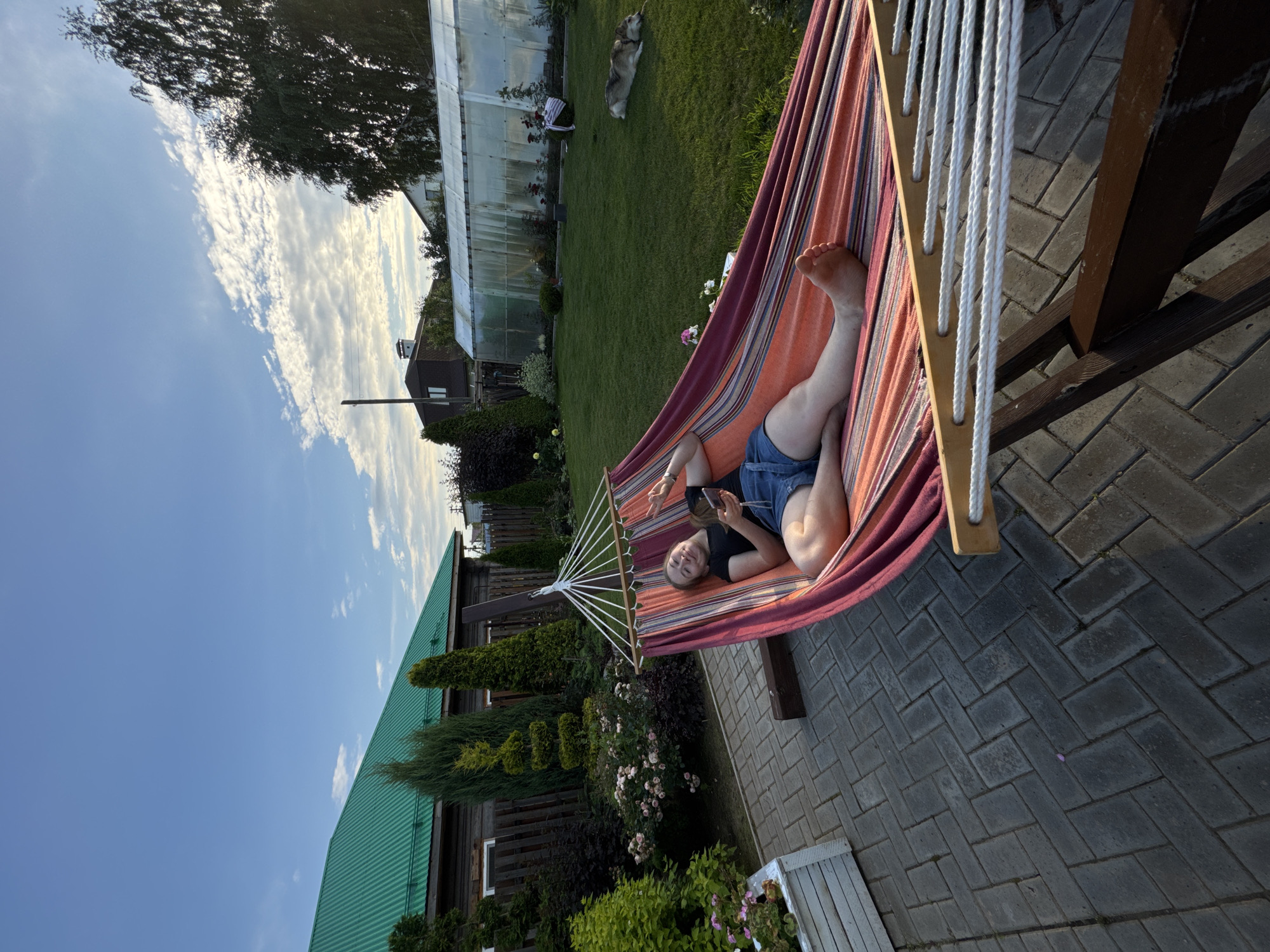 A young woman with blonde hair is lying in a colorful hammock in a backyard. She is wearing a black t-shirt and denim shorts. She is holding a phone in her left hand and has her right hand raised in a peace sign. The hammock is suspended between two wooden posts and is set up on a paved patio. In the background, there is a lush green lawn, a greenhouse, a dog lying on the grass, and a wooden house with a green roof. The sky is blue with scattered white clouds. The location is a backyard in Прикамский, Russia.