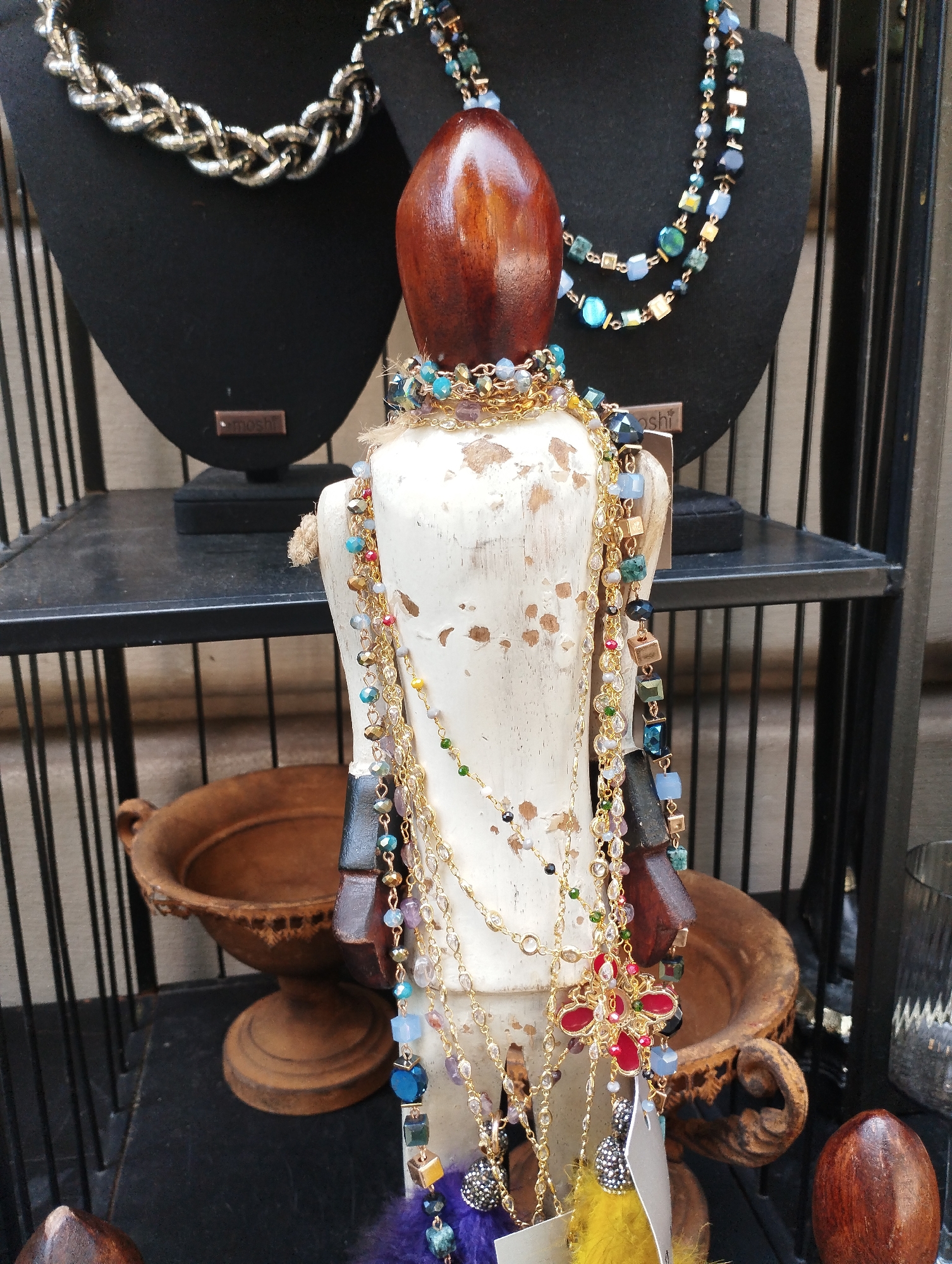 The image shows a retail display of jewelry. In the foreground, a mannequin torso is adorned with multiple layered necklaces. The necklaces are made of gold chains, various beads in shades of blue, green, gold, and purple, and some feature charms. To the left and right of the mannequin, the display shelf holds two decorative, wooden bowls. In the background, two black necklace busts display additional jewelry. The display is enclosed by a black metal grate structure. The scene appears to be set within a shop. There is no discernible indication of the city of Lugano, Switzerland.