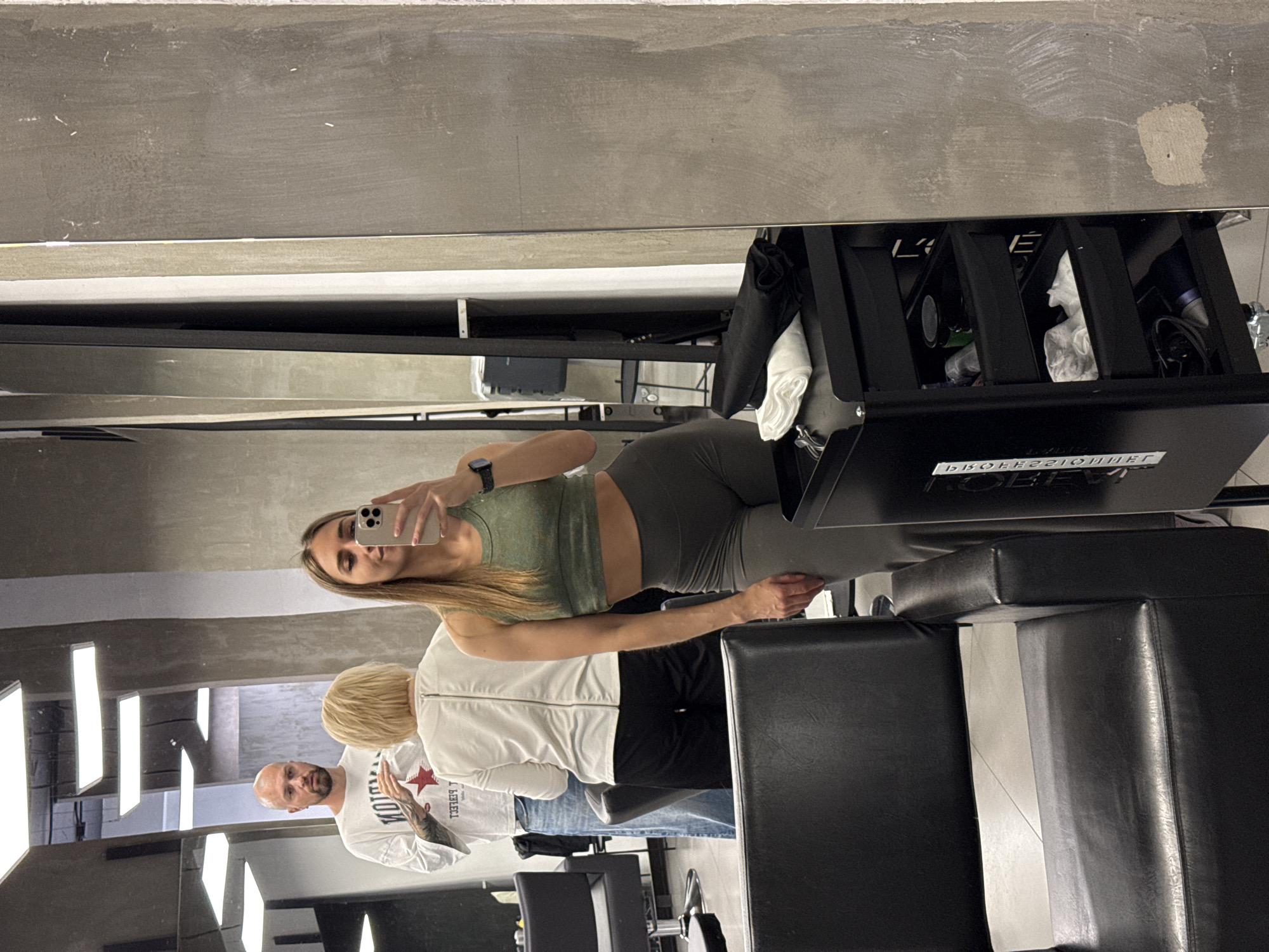 The image appears to be a reflection in a mirror inside a hair salon. A woman wearing athletic clothing (olive green top and leggings) is taking a selfie using her smartphone. Behind her and slightly to her left, a man with a shaved head and a white t-shirt with a red star graphic is visible. An individual with short blonde hair and a white jacket is partially visible between the woman and the man.
Salon equipment, including black styling chairs and a black rolling cart (possibly a stylist's station), are in the foreground. The ceiling of the room features long, vertical light fixtures. The walls are light gray.
Without additional information, it is not possible to conclusively determine if this image was taken in Saint Petersburg, Russia.