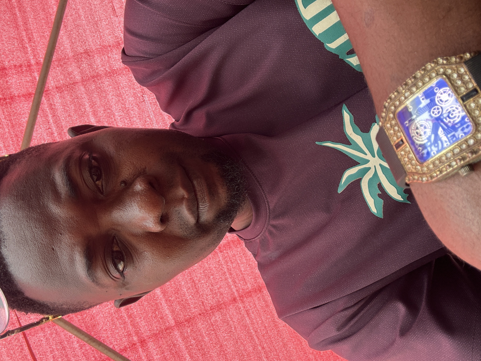 The image is a close-up, high-angle selfie of a Black man in Narayi, Nigeria. He is looking directly at the camera with a neutral expression. He has dark skin, a short beard, and is wearing a maroon t-shirt with a palm tree design. A gold-encrusted watch with a blue digital display is visible on his wrist. The background consists of a textured red surface, possibly a fabric awning, and a thin metal pole. The lighting suggests an outdoor environment.