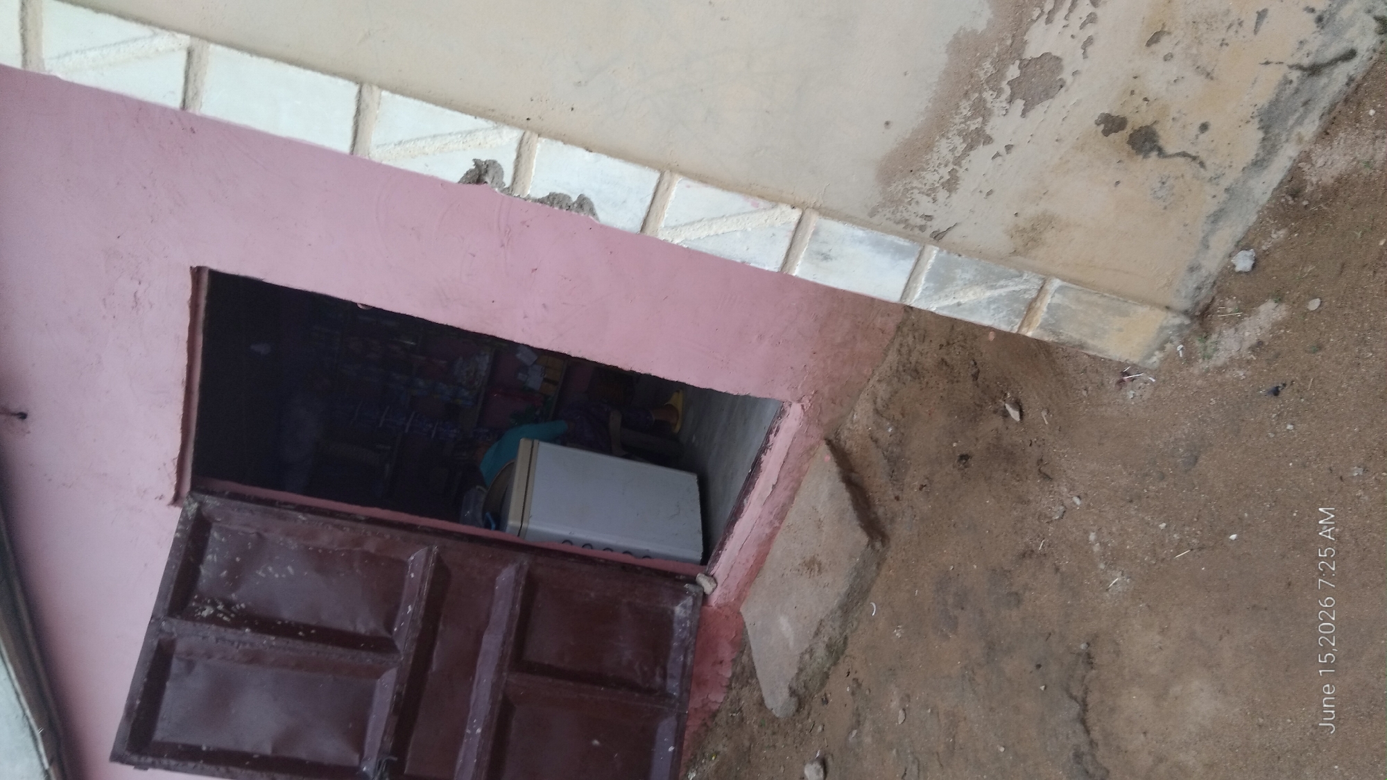 The image depicts a storefront in Howayi, Nigeria, on June 15, 2026, at 7:25 AM. The exterior features a pink-painted wall and a large, dark brown, double door, one panel of which is ajar, revealing the interior. Inside, a white refrigerator is visible. To the right of the doorway, another wall, painted a pale beige and adorned with decorative white rectangular tiles, angles away from the viewer. The ground in front of the store is unpaved, composed of dirt and sand, with a single, rectangular concrete slab serving as a step. The overall scene suggests a typical, modest commercial establishment in a Nigerian town.