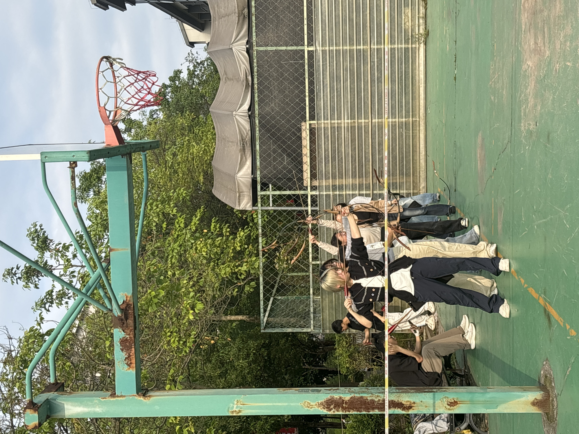 A group of at least eight individuals is practicing archery on a green outdoor basketball court in what appears to be a public park. The location is identified as the city of Jiangjin, China. The court features a basketball hoop with a red and white net, positioned to the left of the archers. A chain-link fence, partially covered by a gray tarp, separates the court from a background of trees. One person in the front row is using a cell phone, while the others are raising their bows. The basketball hoop appears old and rusted. A tall, yellow measuring device is standing close to the group.