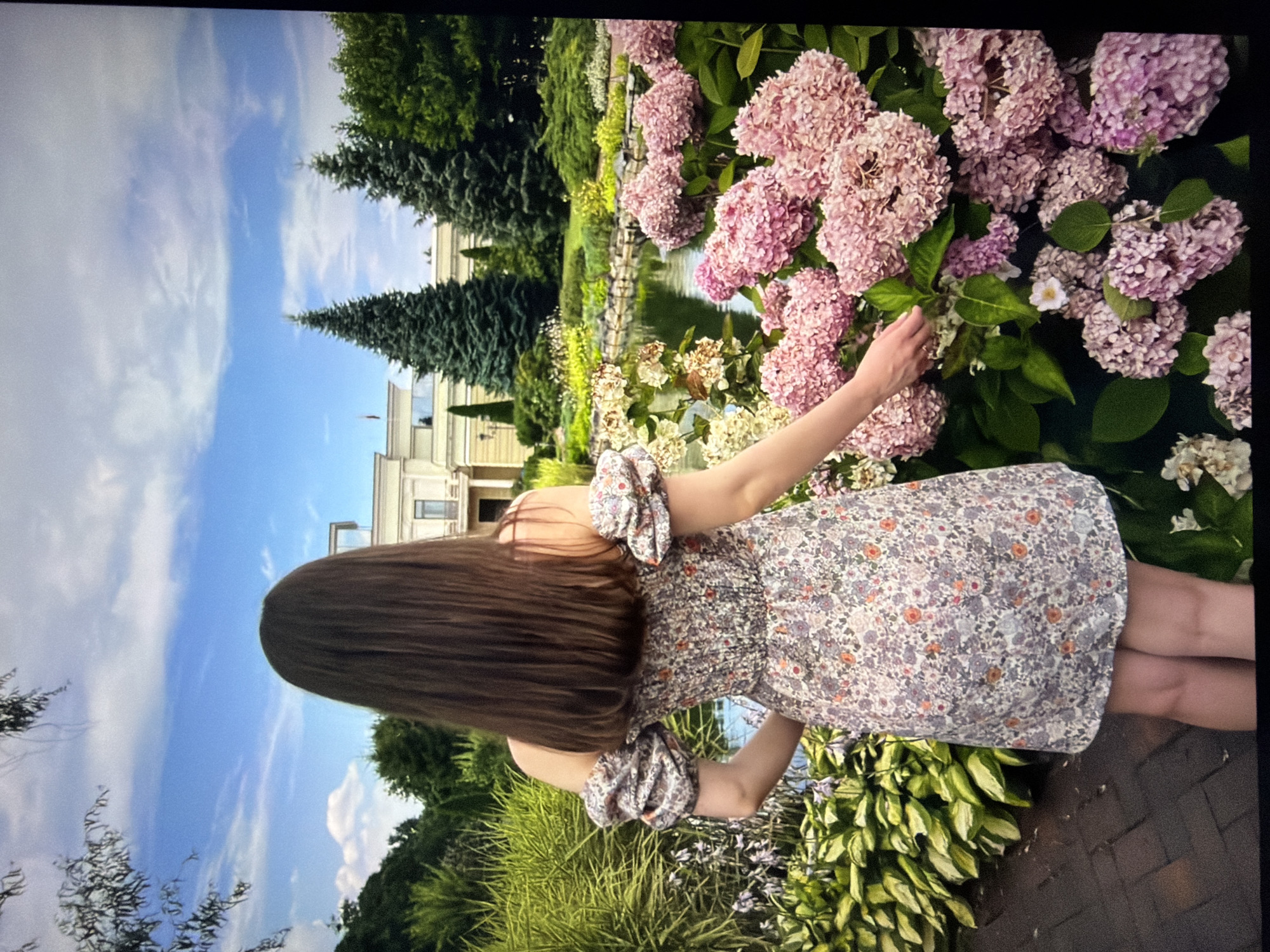 The image depicts a woman in a floral dress standing in a garden with hydrangeas in the foreground. Behind her, a body of water reflects the blue sky and clouds. In the background, a building with classical architecture is visible, surrounded by manicured trees and greenery. The overall scene suggests a leisurely day in a well-maintained park or estate. The location is the National Museum of Ukrainian Decorative Art in Kyiv, Ukraine, a landmark known for its beautiful gardens and serene atmosphere.