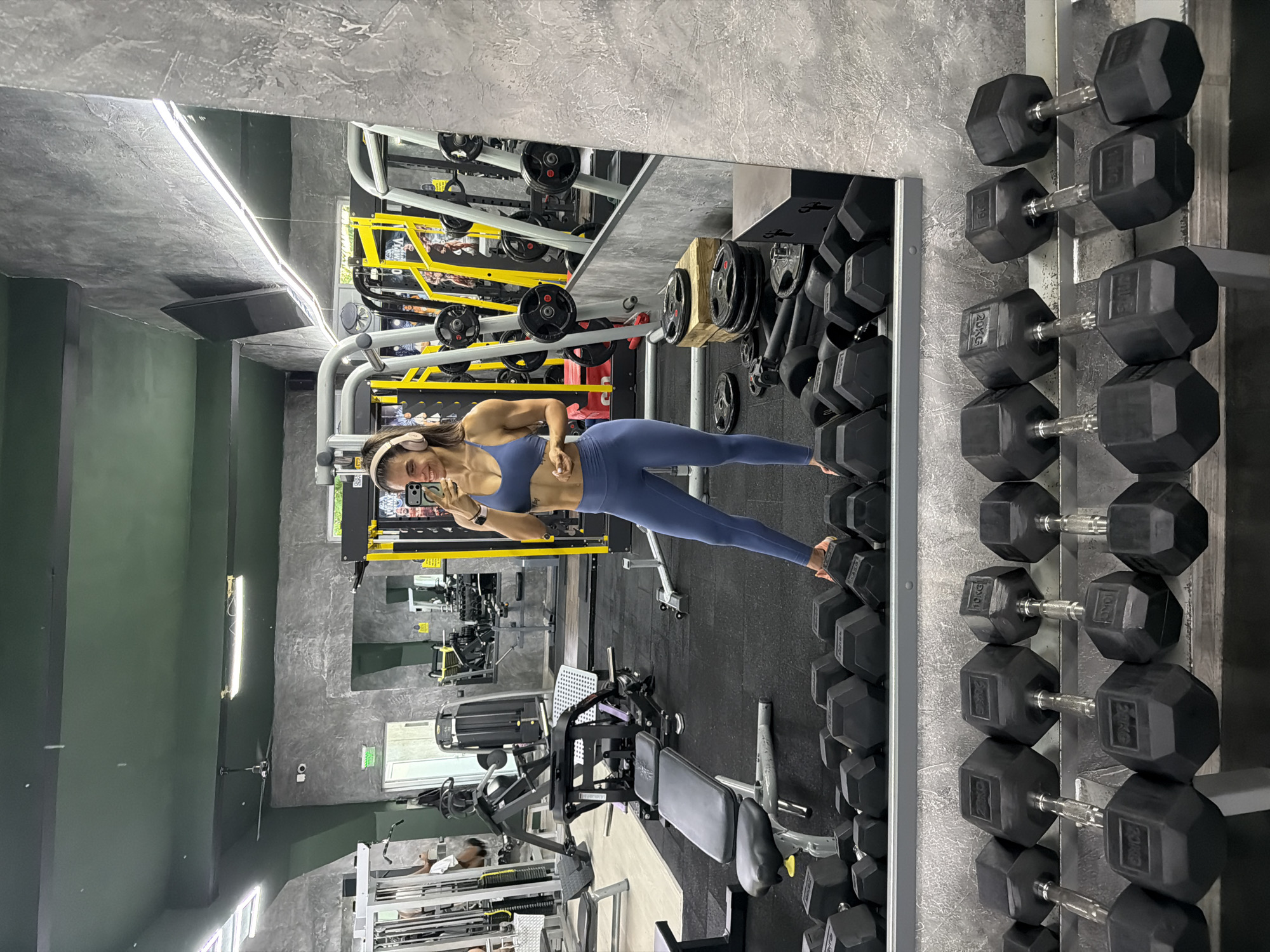 A young woman in athletic attire is taking a selfie in front of a mirror in a gym setting. The gym appears to be well-equipped with various machines, including weight racks holding dumbbells. The background reveals other gym equipment and possibly other individuals exercising. The location is implied to be in Svilengrad, Bulgaria.