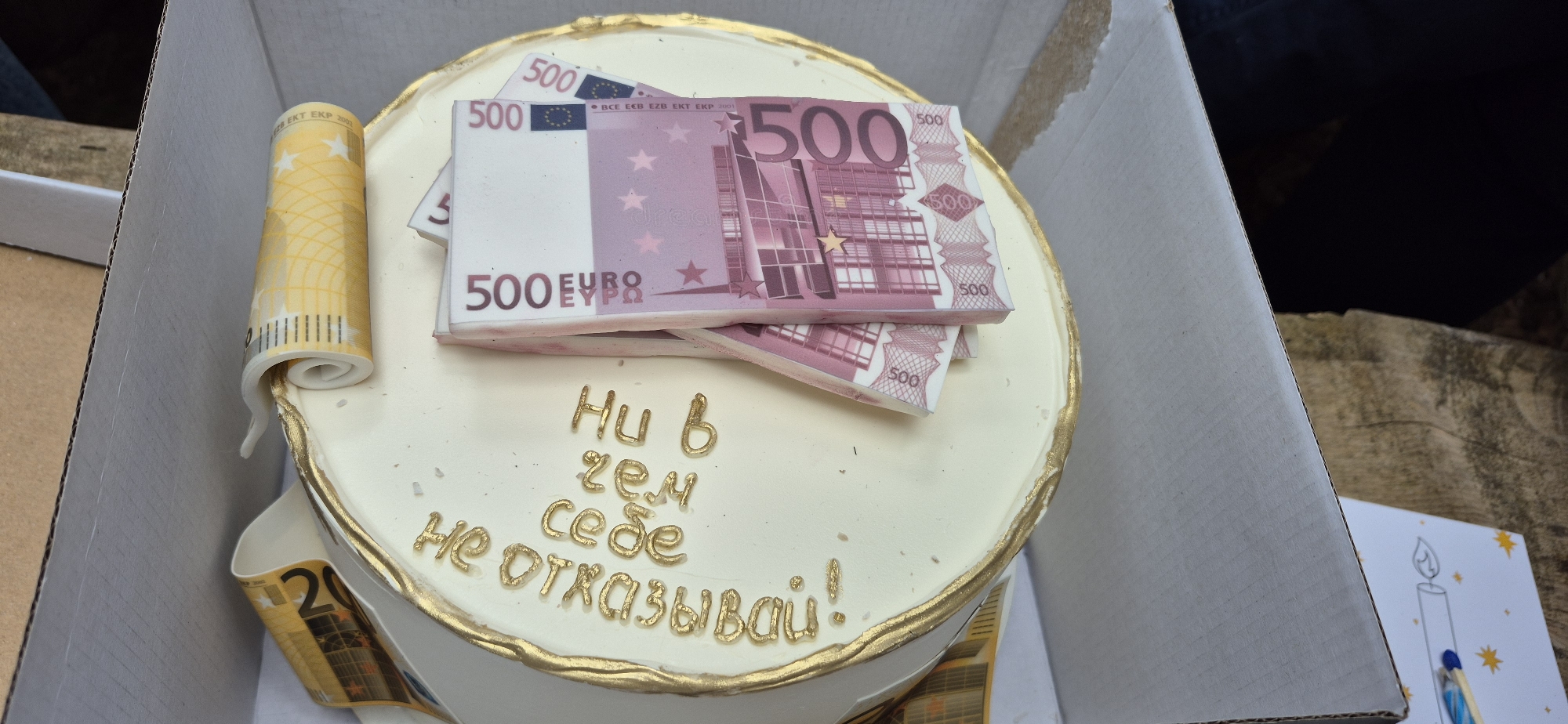 Here is a description of the image:
The image shows a cake decorated with edible replicas of Euro banknotes. There is a stack of 500 Euro notes on top of the cake and rolled-up 20 Euro notes around the base. The cake is white with a gold trim and has the text "Ни в чем себе не отказывай!" written on it in gold icing. The cake is in a cardboard box, and a greeting card with a candle drawn on it is visible next to the box. The image was taken indoors. There are no people visible. The location, Mali Bostur, Montenegro, cannot be confirmed based on the visual information.