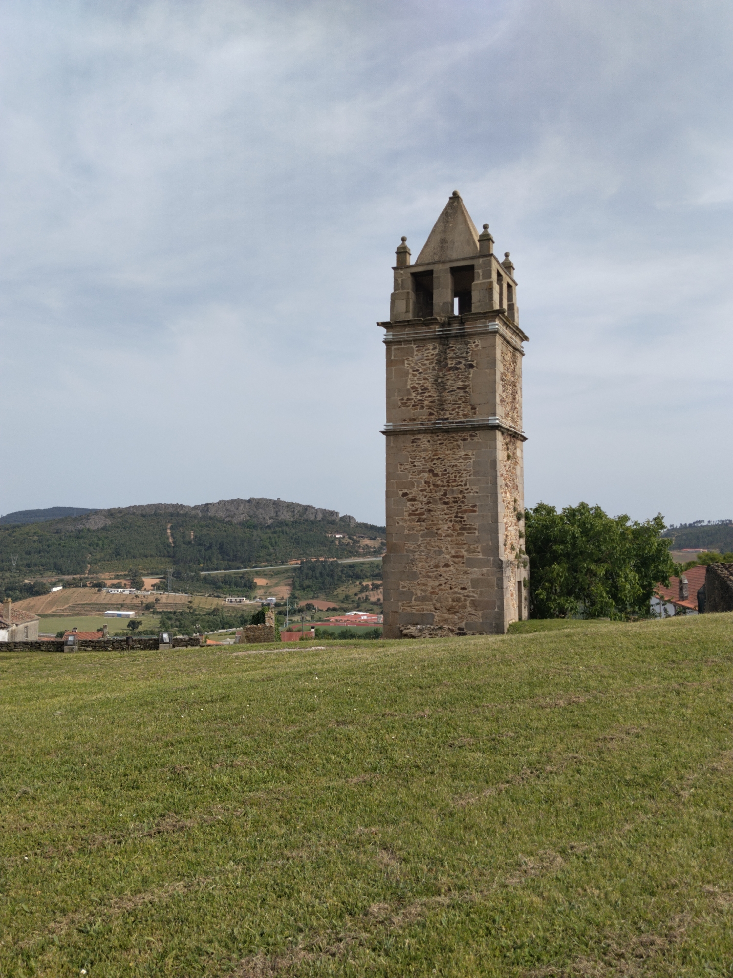 The image shows a tall, stone bell tower located in Mogadouro, Portugal. The tower is the main subject and is composed of two primary sections, the lower one being wider and constructed with rough-hewn stones, while the upper section is narrower with smooth stone corners and features open archways at the top, topped with a pyramidal roof. The tower stands on a grassy hill that slopes down towards the foreground. Behind the tower, there are trees and rooftops that suggest a small village. In the background, rolling hills and mountains covered in forests are visible under a cloudy sky.