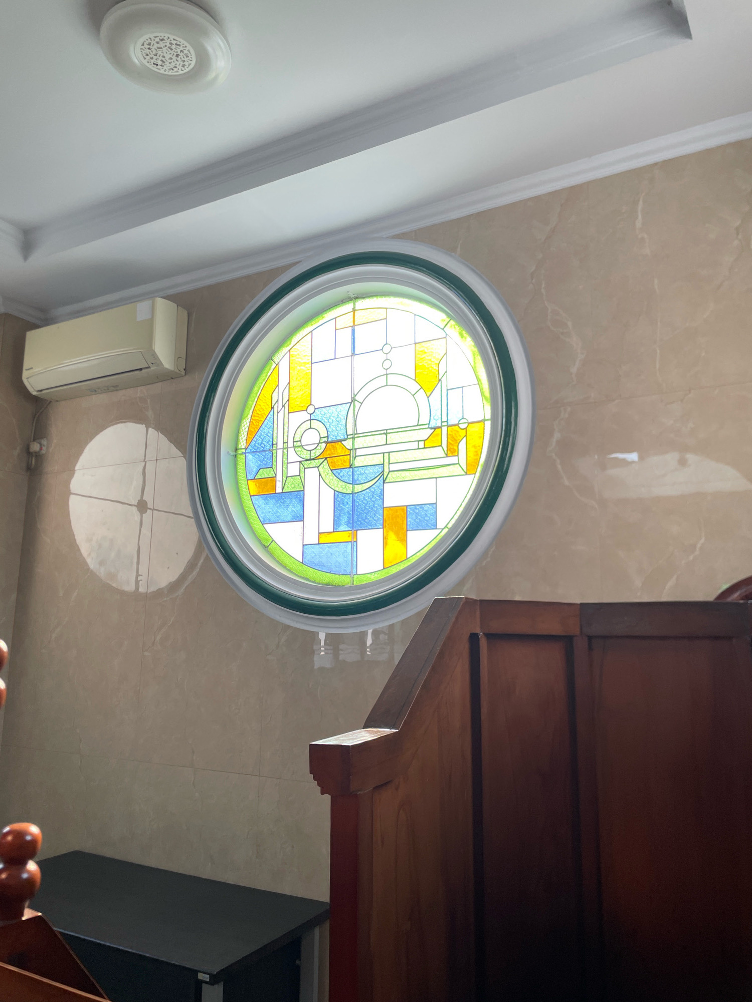 The image shows a circular stained-glass window with a geometric design featuring yellow, blue, and white panels, set against a beige marble-patterned wall. Above the window, a white ceiling with decorative molding is visible, along with a white ceiling light fixture and an air conditioning unit mounted on the wall. To the right of the window, a portion of a dark wooden structure, possibly a pew or part of a pulpit, is in the foreground. To the left of the window, a circular shadow with a cross pattern is cast on the wall, likely from a light source. A dark rectangular object, possibly a cabinet or desk, is partially visible at the bottom left of the image. The scene appears to be indoors, possibly in a place of worship or a room with decorative architectural elements. The lighting suggests it is daytime. The general aesthetic suggests a relatively modern building with some traditional decorative touches. The specific location within Tangerang, Indonesia, cannot be determined from the image alone.
