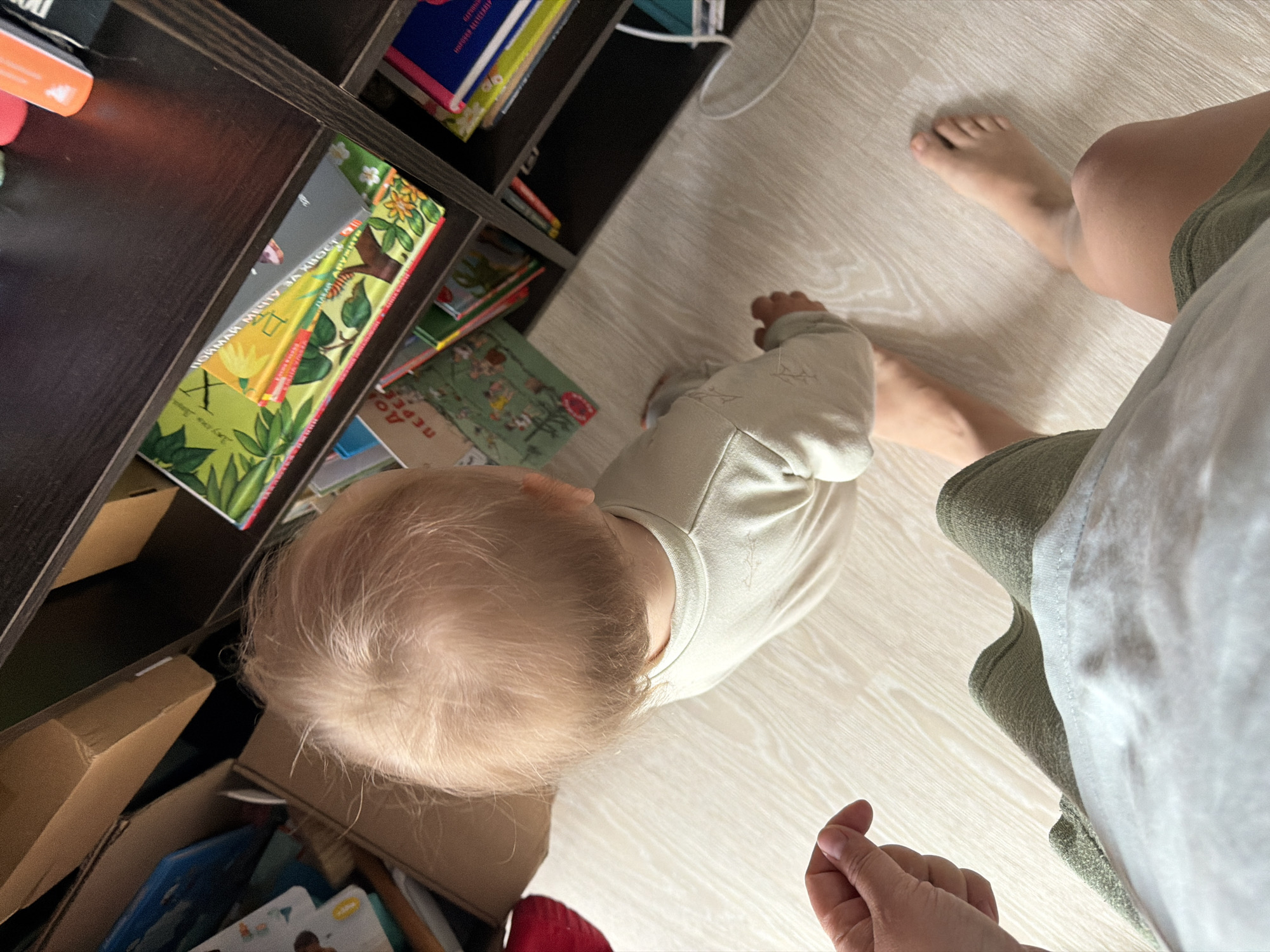 The image shows a partial view of a room with a light-colored wooden floor. A dark brown bookshelf filled with books and some boxes is visible on the left side of the frame. A small child with blond hair, wearing a light-colored outfit, is standing near the bookshelf, looking towards the lower shelves. A person's legs and bare foot are visible on the right, along with part of their clothing. The person's hand is also visible at the bottom. The location is Chaikovsky, Russia, but there is no identifiable element in the image to directly confirm this.