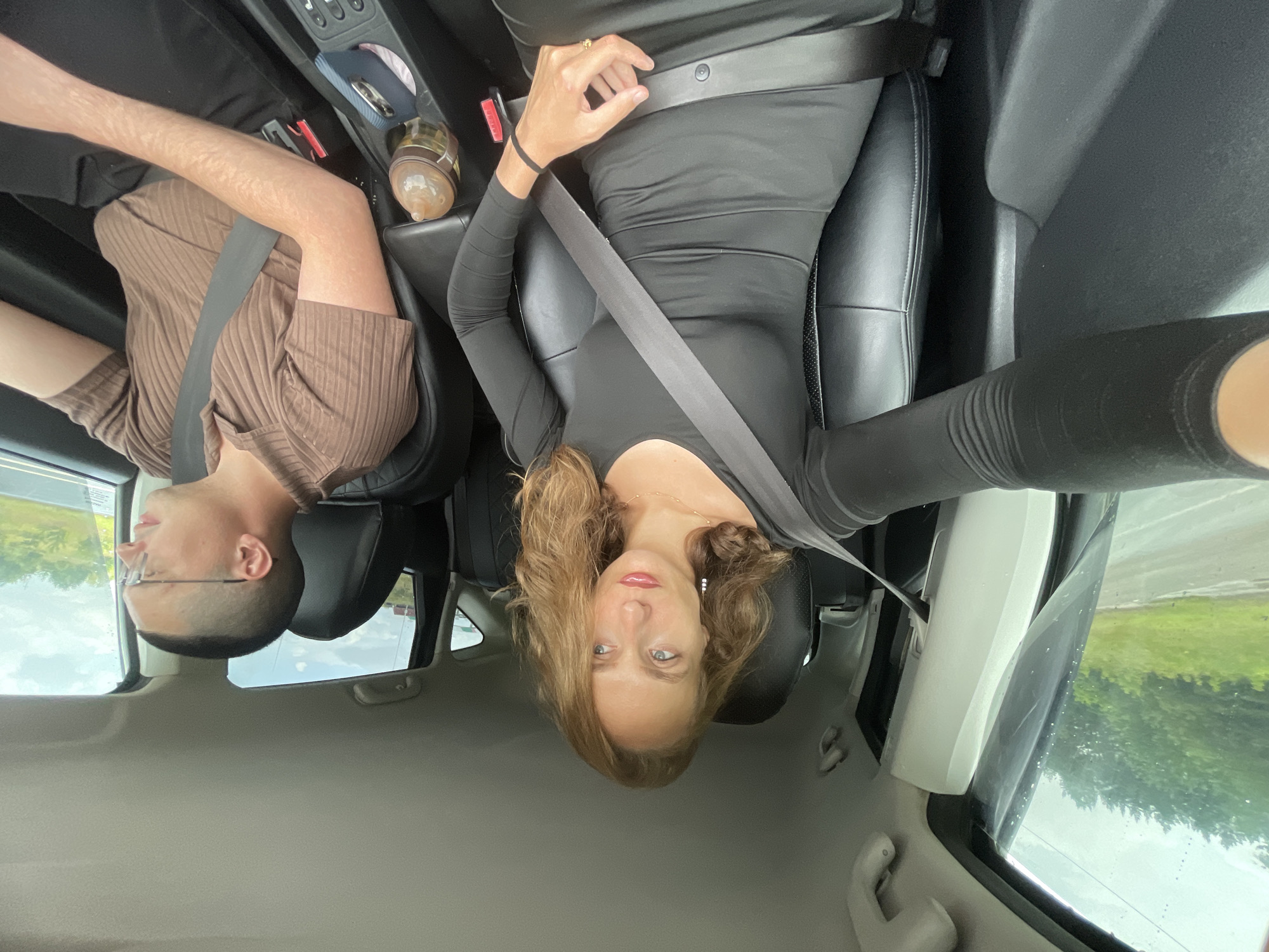 A medium shot shows two individuals inside a car, both wearing seatbelts. The woman in the foreground, with shoulder-length blonde hair and red lipstick, is looking to her right, adjusting her seatbelt. She is wearing a form-fitting black long-sleeved dress. To her right, in the driver's seat, a man with a buzz cut and glasses is looking forward and slightly to his right. He is wearing a brown ribbed polo shirt and black pants. The car's interior is visible, with black leather seats and the central console. Outside the windows, a blurred green landscape with trees and a cloudy sky suggests they are traveling on a road, possibly on the outskirts of Shud'ya, Russia.