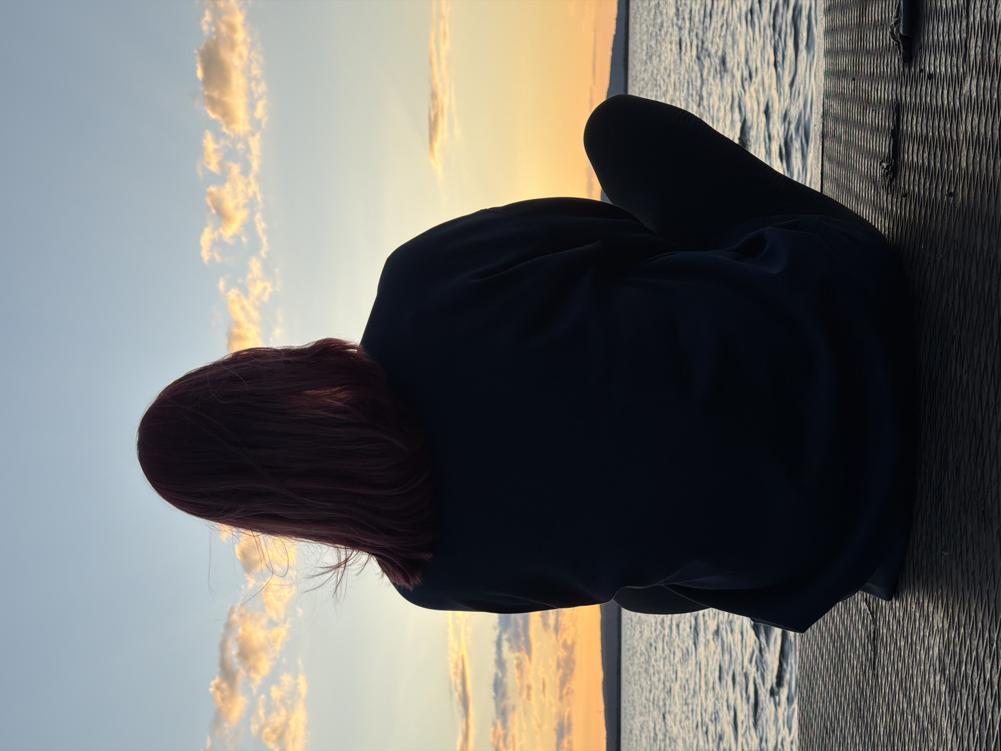 A person with reddish-brown hair, viewed from behind, sits facing a body of water at sunset. They are wearing a dark-colored sweatshirt and dark leggings, with their legs crossed. The person is seated on a dark, textured surface, likely a pier or boardwalk. The sun is low on the horizon, casting a warm orange and yellow glow across the sky, with scattered clouds tinged with pink and orange. The water is calm, reflecting the colors of the sunset. A faint silhouette of land can be seen in the distance. The location is not specified in the image itself, and therefore cannot be identified as Chaikovsky, Russia.