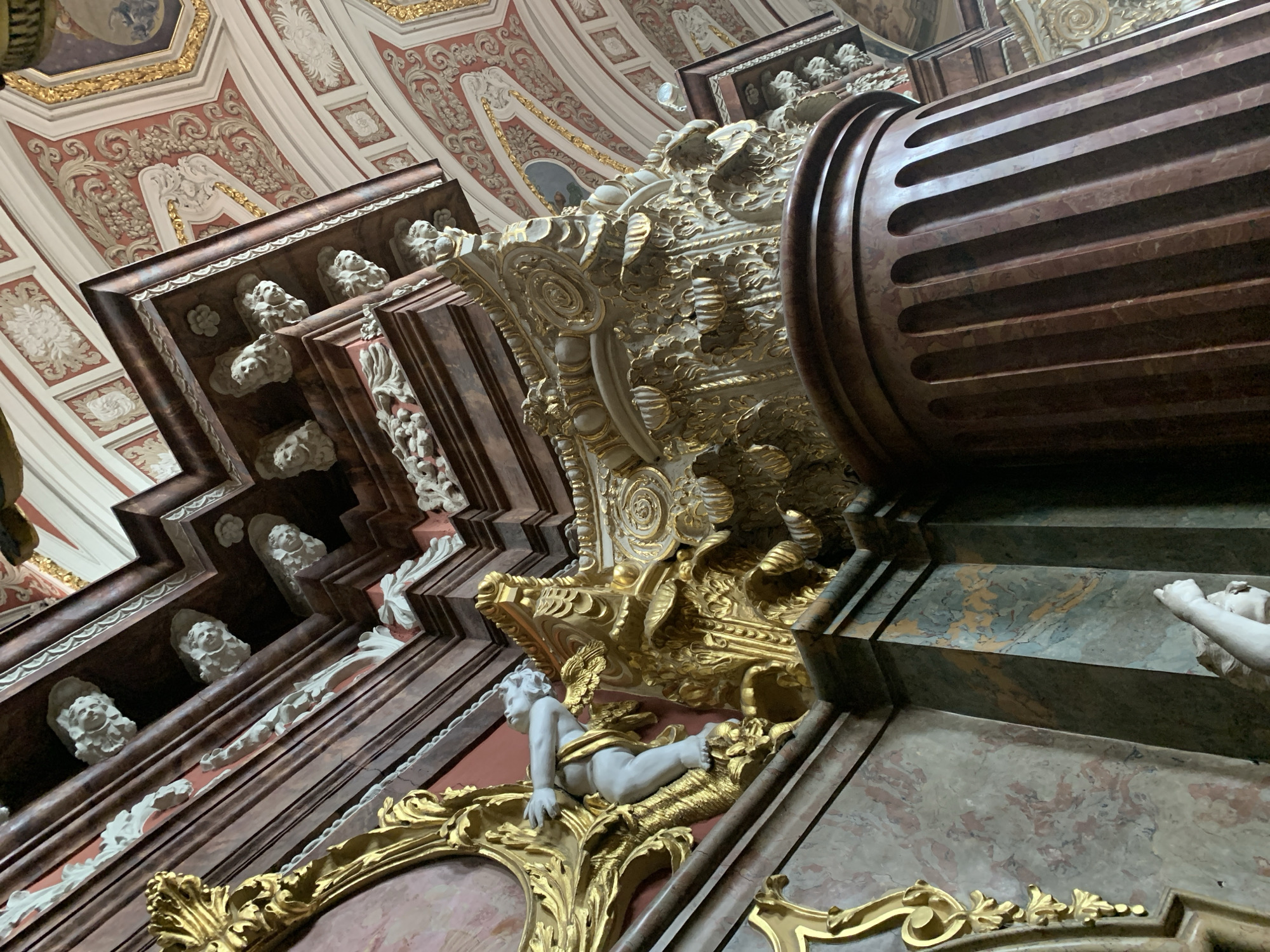 This image captures an ornate interior, likely a church or historical building, featuring a large, fluted column made of reddish-brown marble. The column is topped with elaborate golden ornamentation that cascades downwards, resembling foliage and scrolls. Above this, a segment of a richly decorated ceiling is visible, painted with intricate patterns in red, gold, and white.
On the left side of the image, a golden, Baroque-style architectural element frames a white statue of a cherub or angel. The cherub is depicted with outstretched arms, appearing to support or interact with the golden decorative work. Further down the wall, another statue of a cherub's arm is visible, reaching upwards. The walls themselves are adorned with marble-like patterns in shades of pink, grey, and green.
The style of the architecture and decoration suggests it is part of a significant Baroque or Rococo interior. Considering the context of Poznan, Poland, this scene is highly reminiscent of the interiors found in the Archcathedral Basilica of St. Peter and St. Paul, particularly elements within its chapels. The detailed carvings, gilded stucco, and rich marble work are characteristic of Baroque artistry commonly found in prominent religious sites in European cities. The detailed ornamentation suggests a high level of craftsmanship and historical significance.